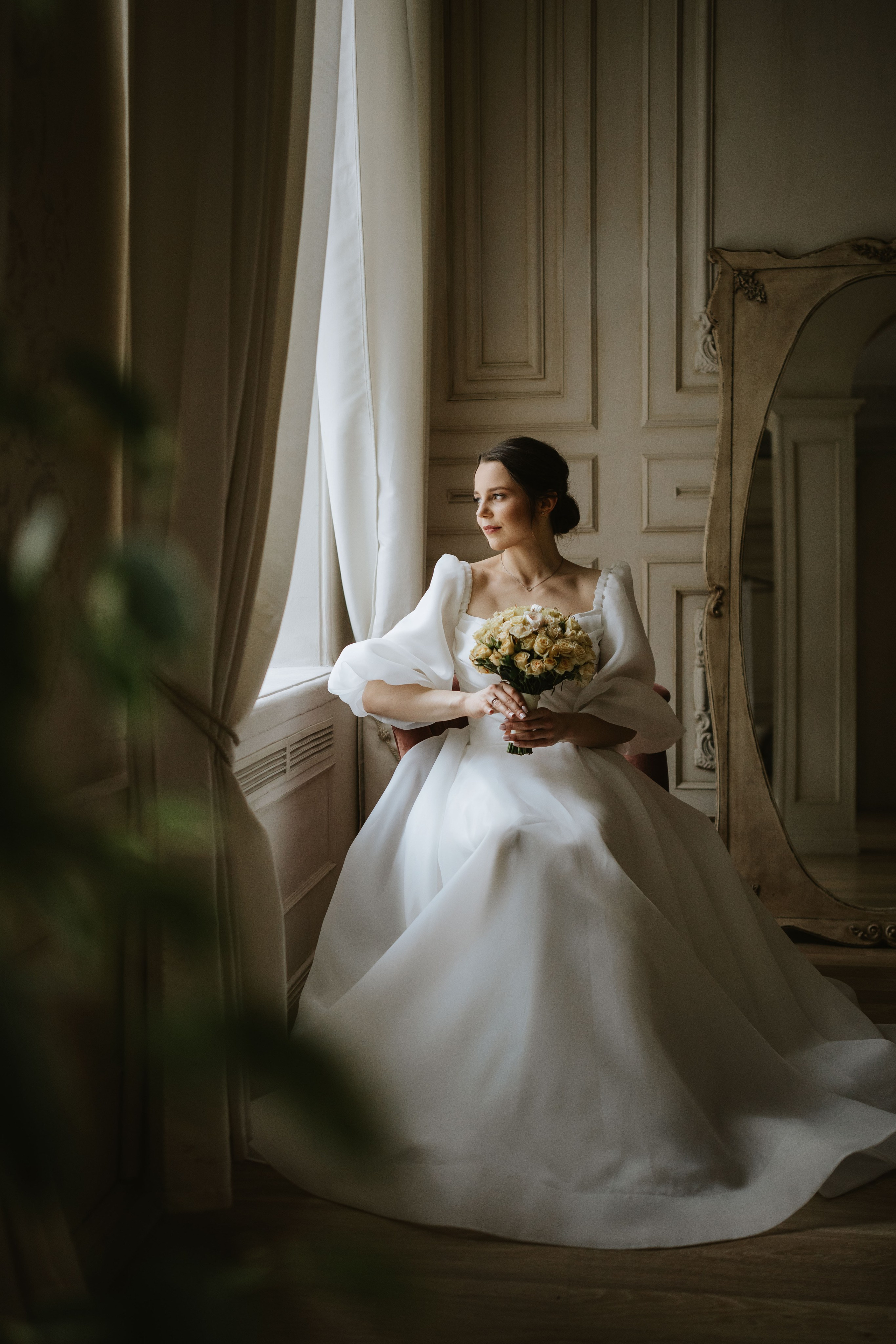 “Una sposa seduta accanto a una grande finestra in una stanza elegante, che guarda fuori con un'espressione serena. Indossa un abito da sposa bianco con maniche a sbuffo e tiene in mano un bouquet di rose beige. Uno specchio a figura intera riflette parte della stanza alle sue spalle e la luce soffusa proveniente dalla finestra crea un'atmosfera sognante.”