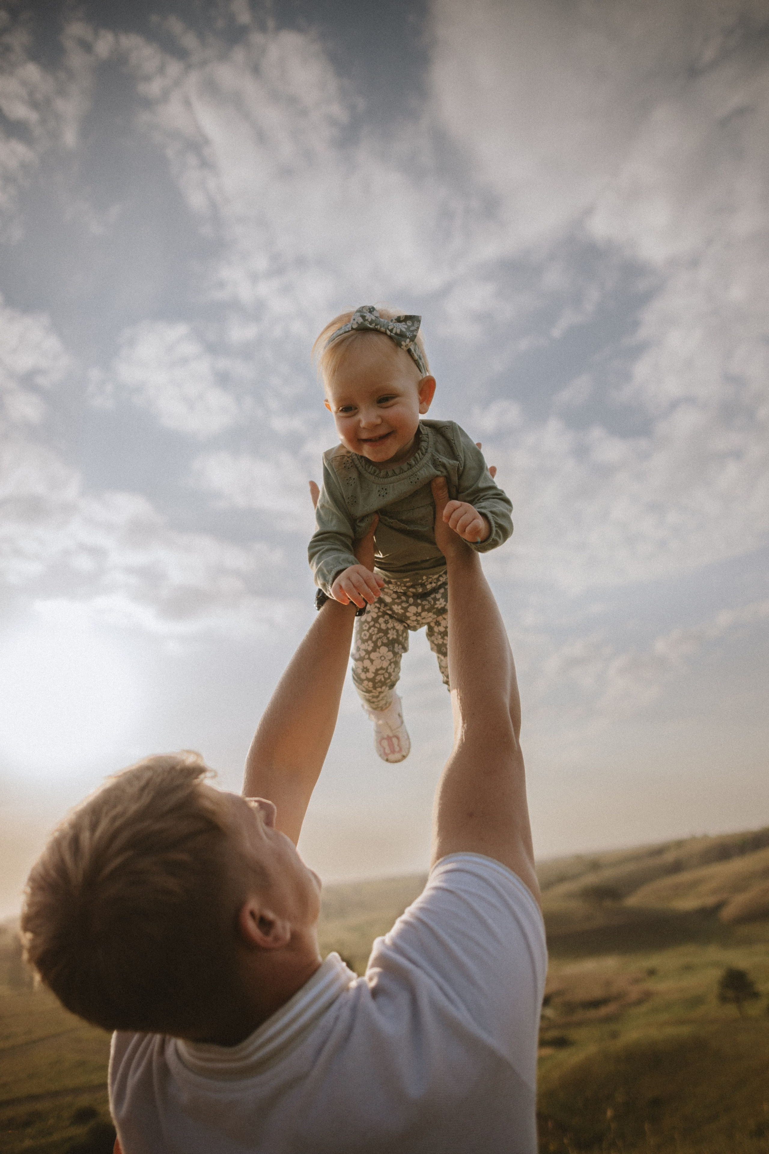 “Una bambina gioiosa con una fascia in testa che viene sorretta dal padre in un ambiente esterno soleggiato. La bambina sorride e si gode il momento mentre il padre la solleva sullo sfondo di un cielo luminoso e di dolci colline.”