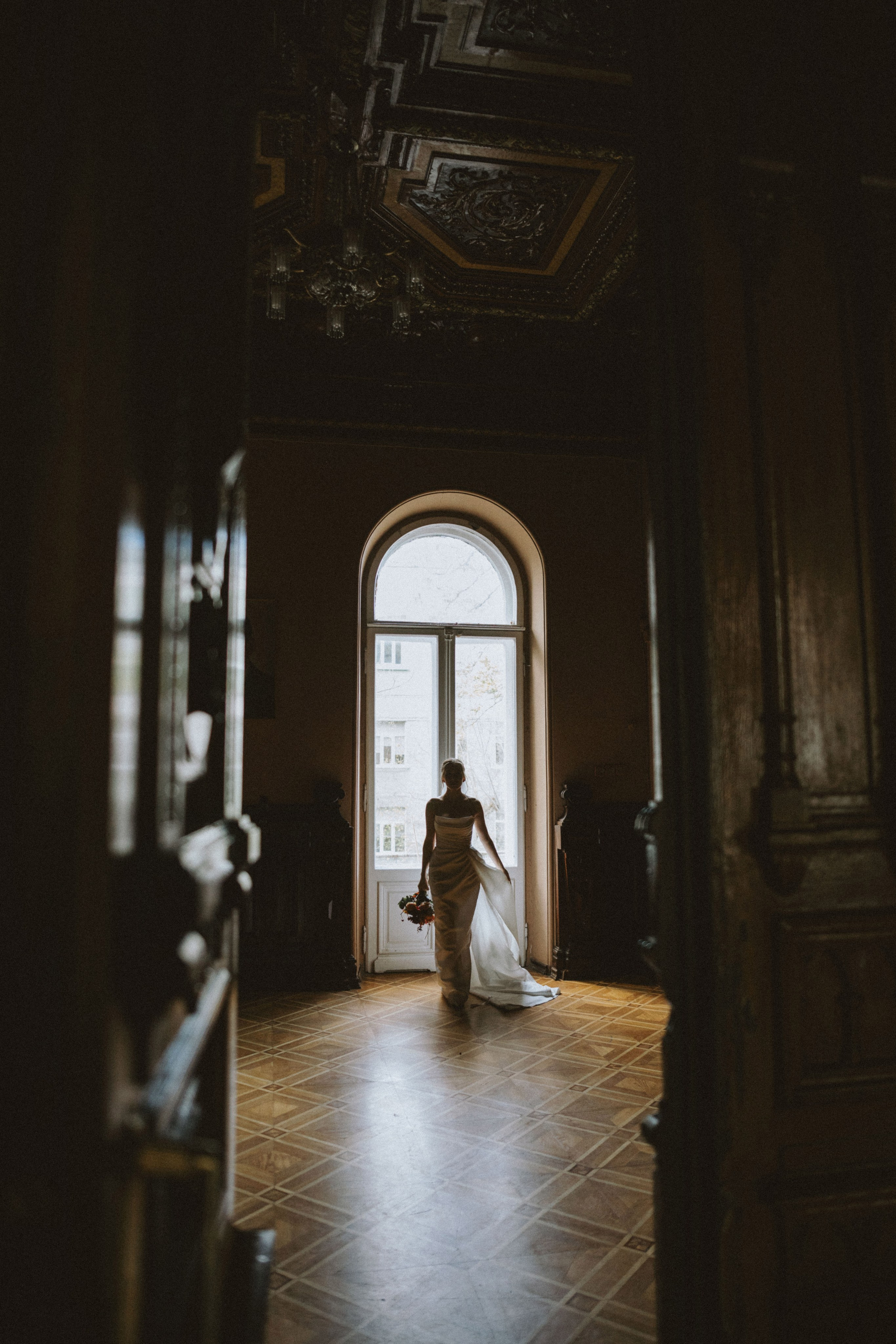 “Una sposa in uno splendido abito da sposa, con in mano un bouquet, cammina verso una grande finestra. La stanza è poco illuminata, con splendidi dettagli in legno sulle cornici delle porte, che creano un effetto drammatico e di silhouette quando la sposa entra nella luce”.