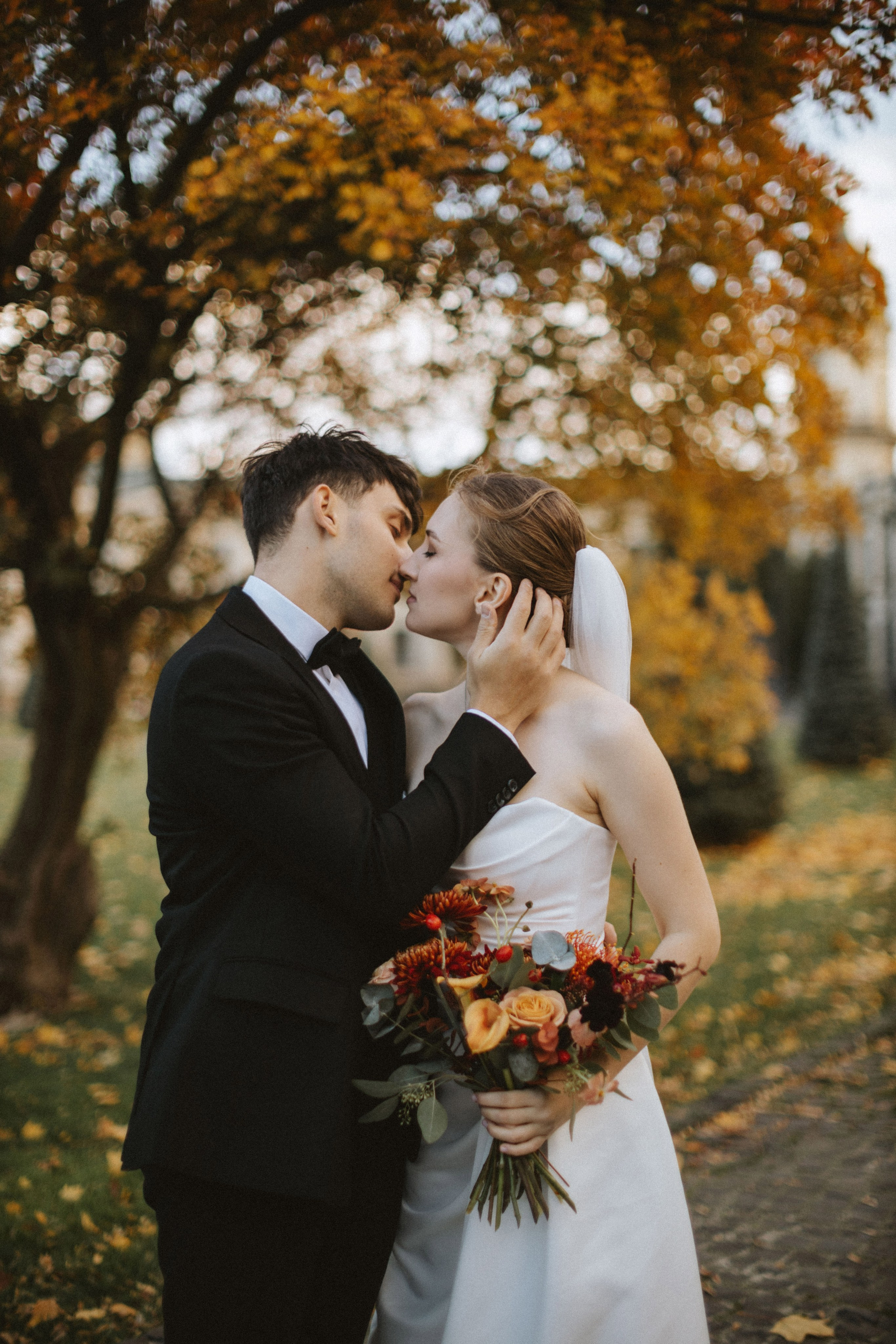 “La sposa e lo sposo si scambiano un tenero bacio tra il vibrante fogliame autunnale. La sposa tiene in mano un bouquet di fiori dai toni caldi, mentre lo sfondo di foglie dorate e una scena morbida e sfocata creano un momento romantico perfetto per il loro matrimonio autunnale.”