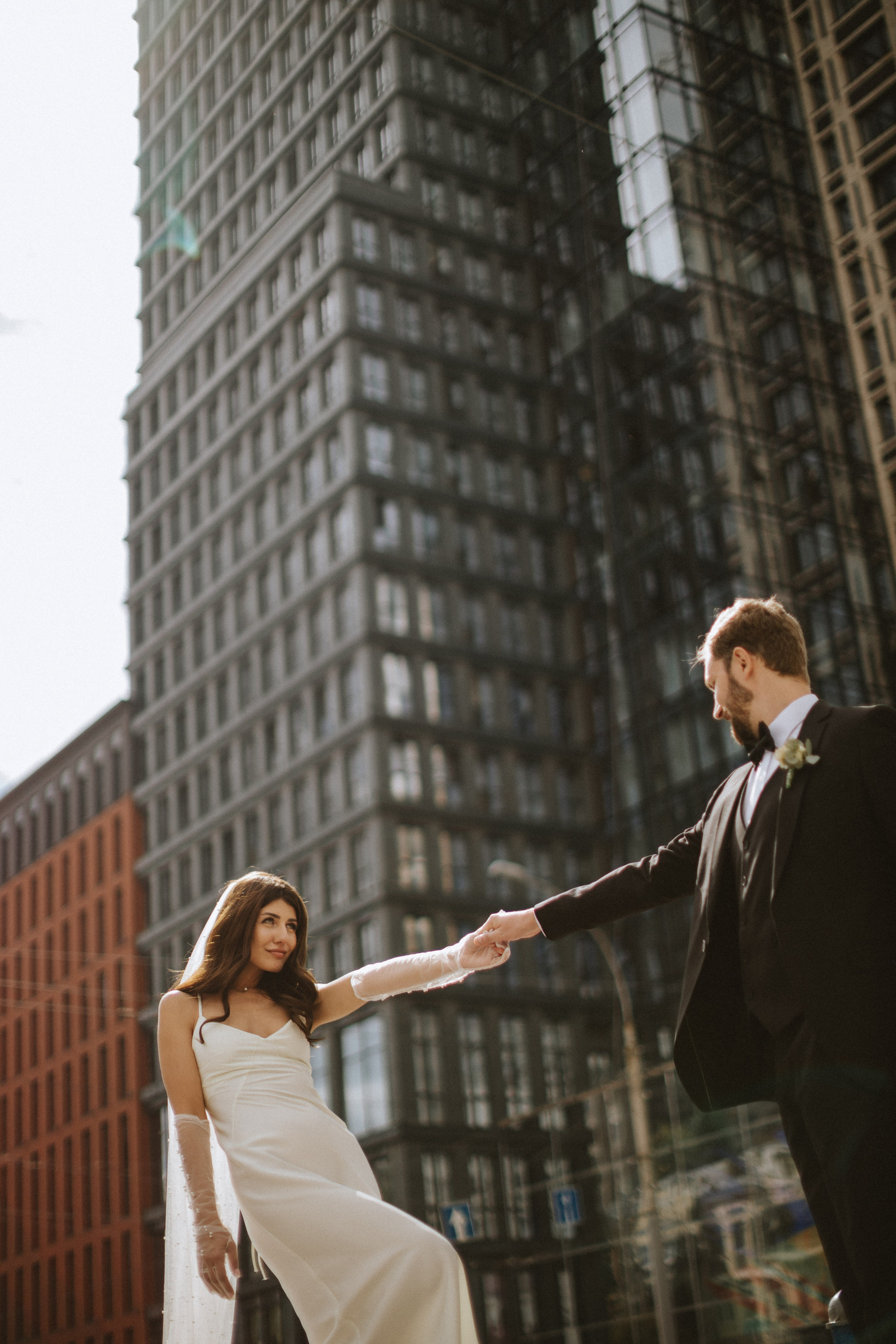“Una coppia si trova all'esterno di un alto edificio, immersa nella calda luce del sole. La sposa, con un elegante abito bianco e un velo, tiene la mano dello sposo e gli sorride. Lo sposo, vestito con uno smoking nero con papillon, la tira dolcemente verso di sé. Il paesaggio urbano moderno li circonda, con i vetri riflettenti degli edifici che creano un elegante sfondo urbano”.