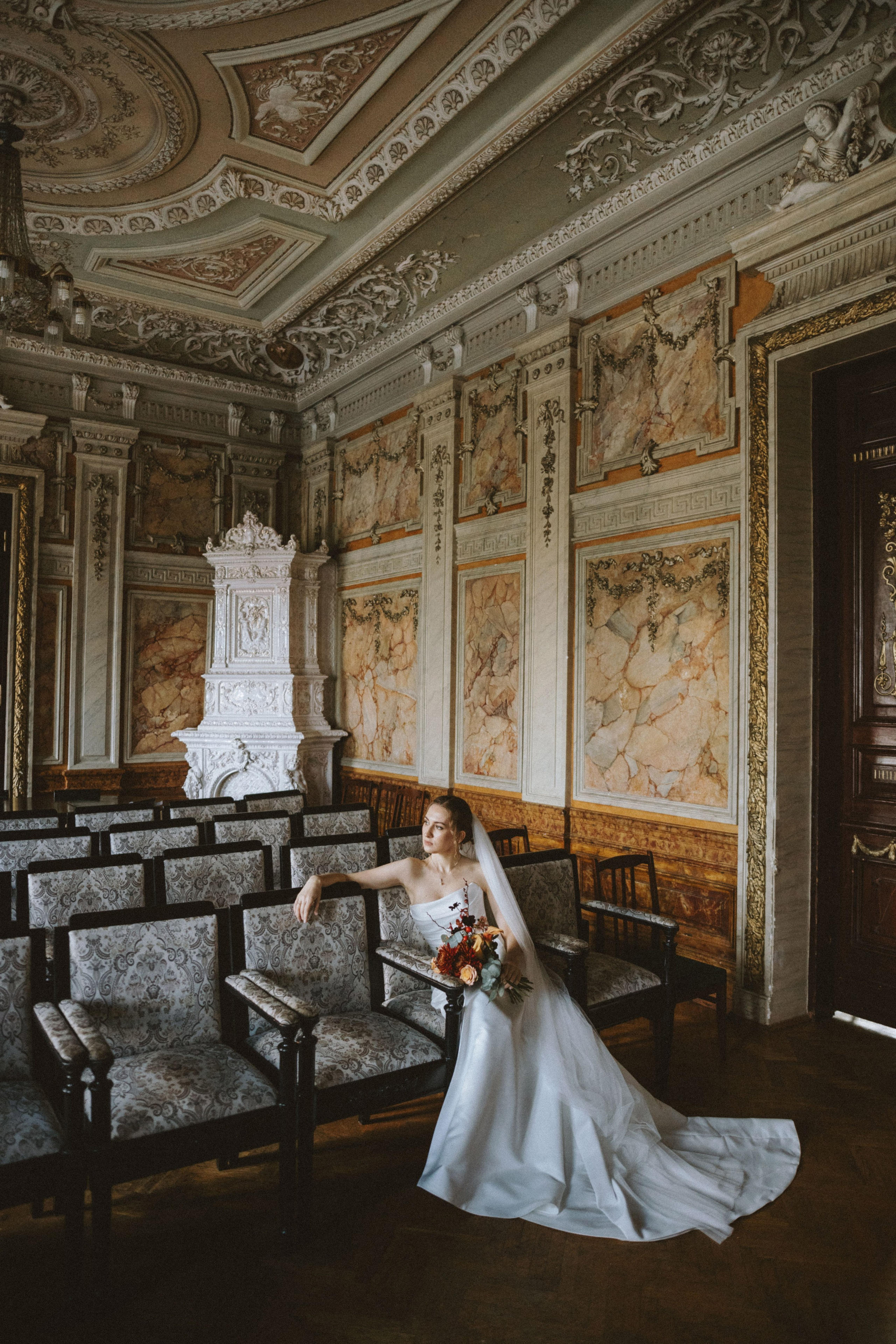 “La sposa in una lussuosa sala in stile vintage, seduta su una sedia con un bouquet floreale in mano. L'interno elegante presenta pareti intricate, mobili classici e un grande camino”.