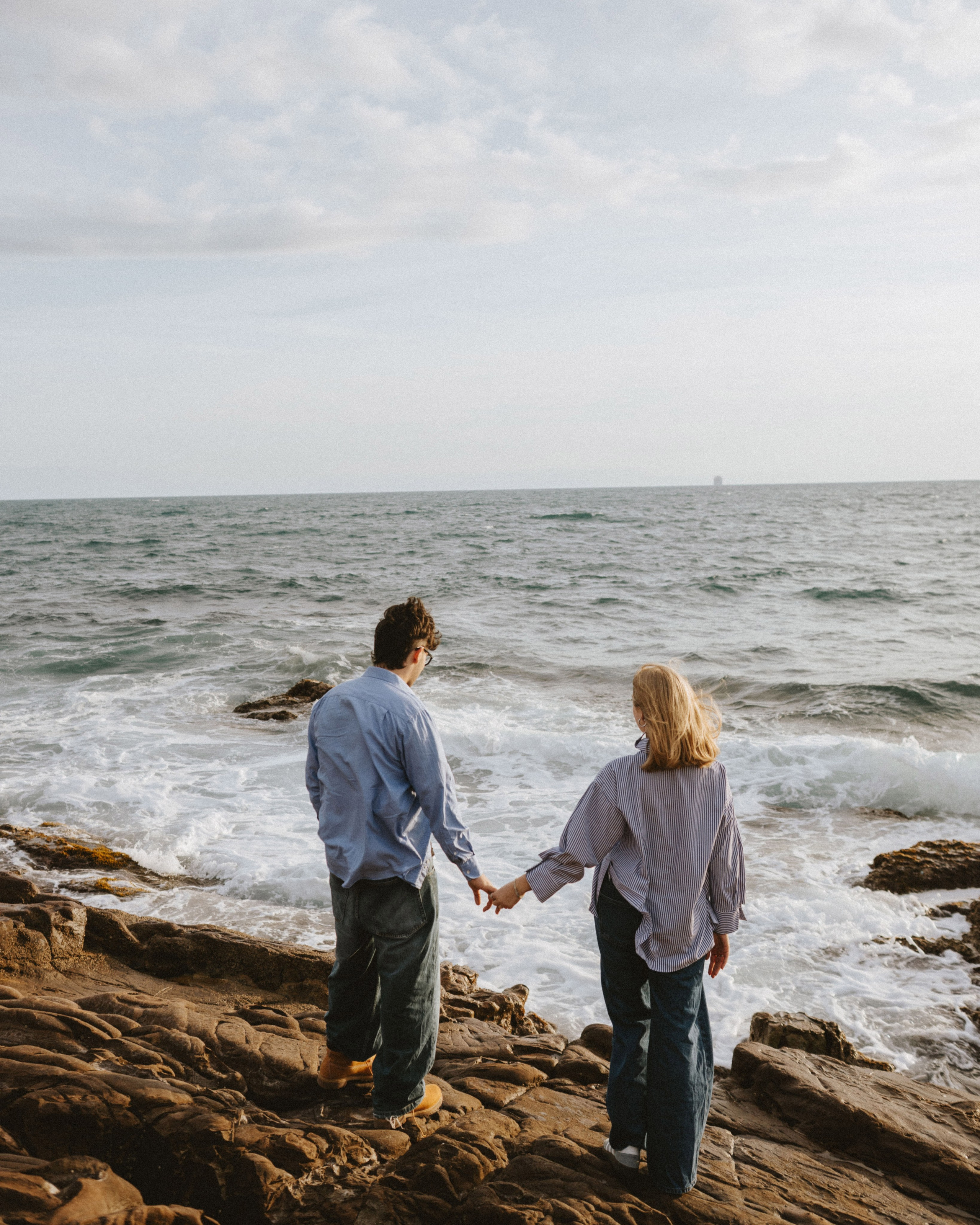 "Romantic couple photoshoot by the sea near Livorno, Italy. A man and a woman hold hands on rocky cliffs with ocean waves in the background. Natural and emotional couple photography."
