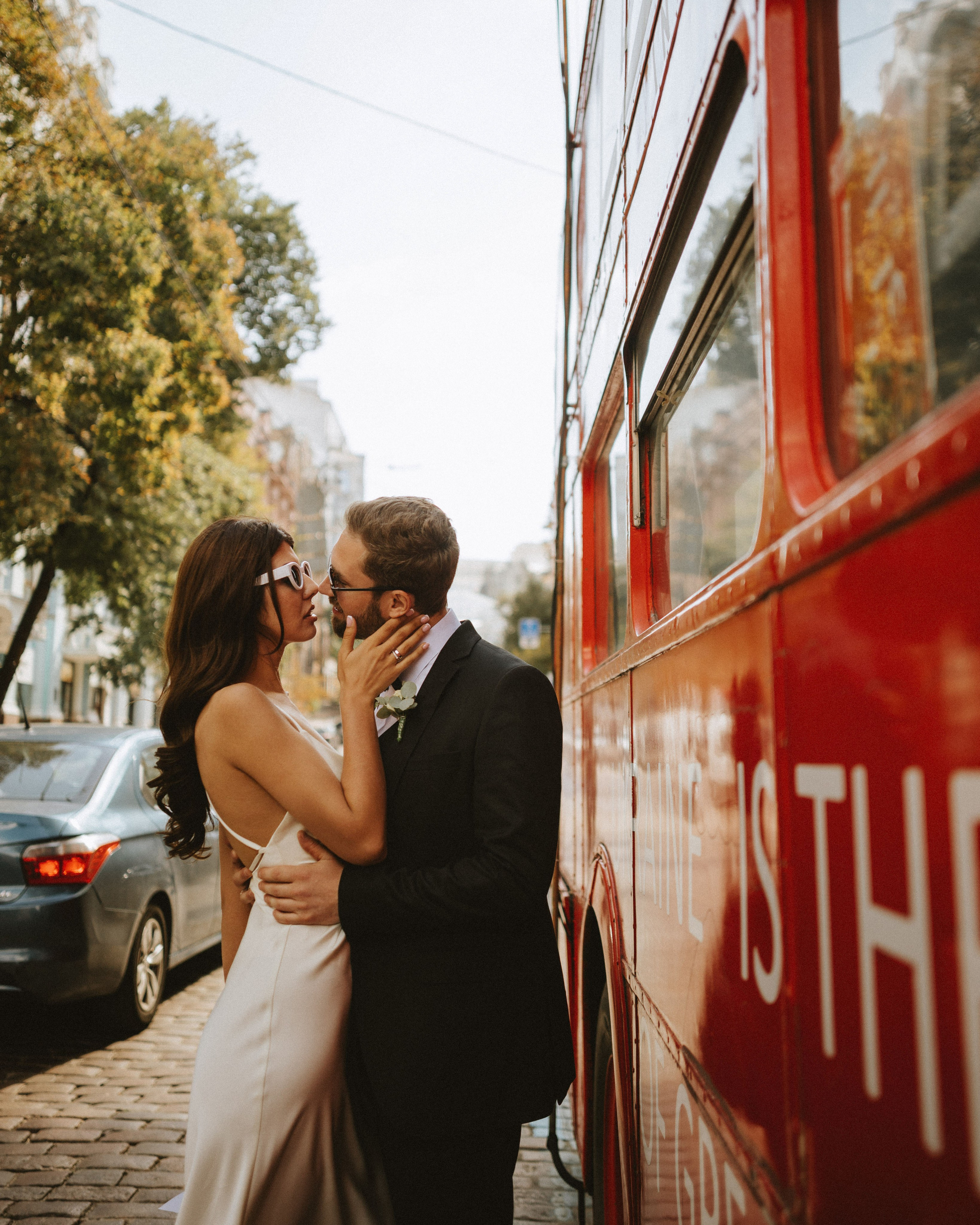 “Una coppia di sposi è vicina e si bacia accanto a un autobus a due piani di colore rosso vivo. La sposa, che indossa un elegante abito bianco senza schienale e occhiali da sole, abbraccia lo sposo, che indossa un formale abito nero con camicia e cravatta bianca. L'ambientazione urbana e l'illuminazione calda creano un'atmosfera romantica e moderna”.