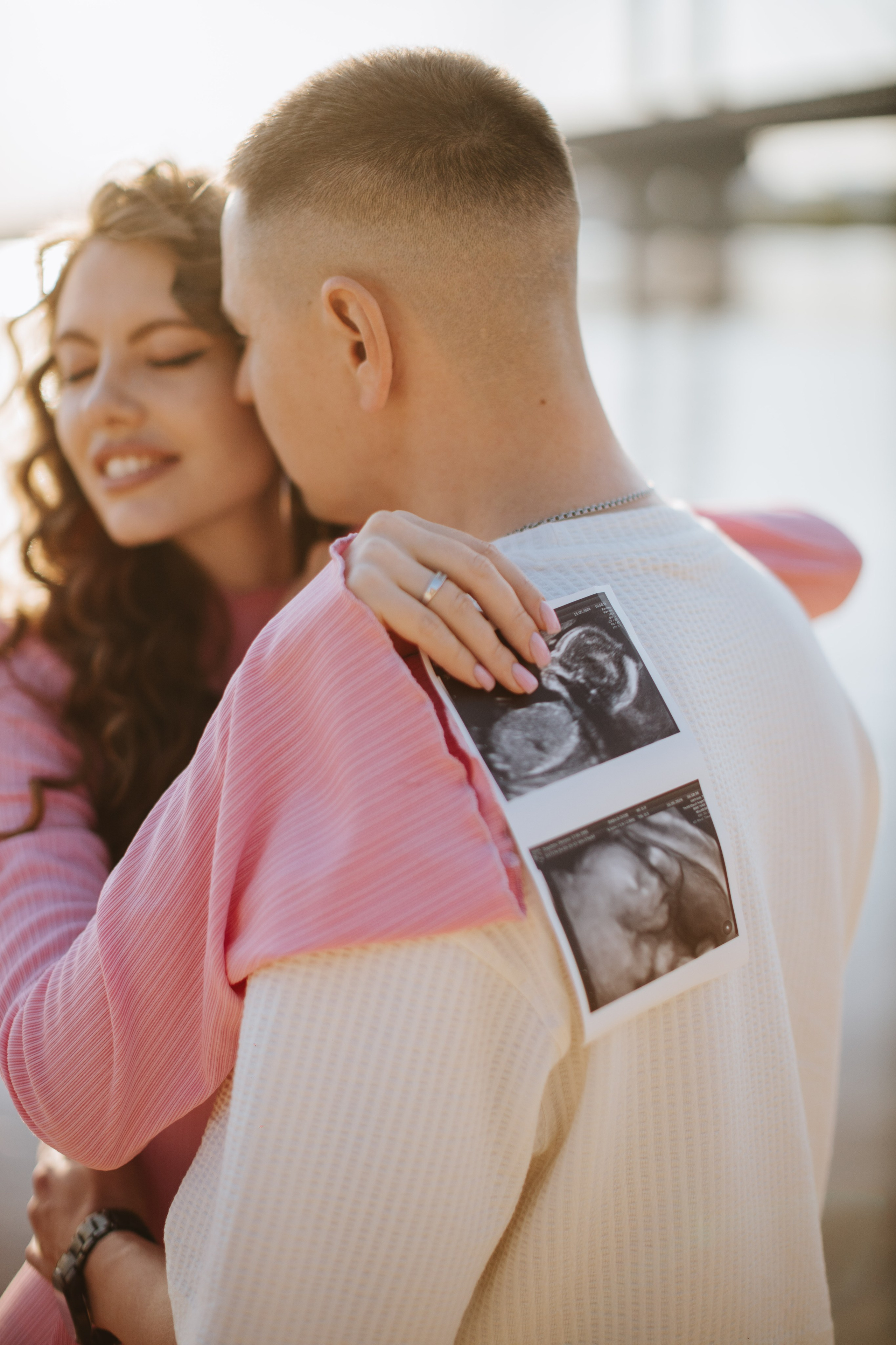 “Una coppia condivide un momento di tenerezza con la donna che tiene le foto dell'ecografia sulla schiena dell'uomo. La coppia si trova all'aperto e la luce del sole splende dolcemente su di loro”.