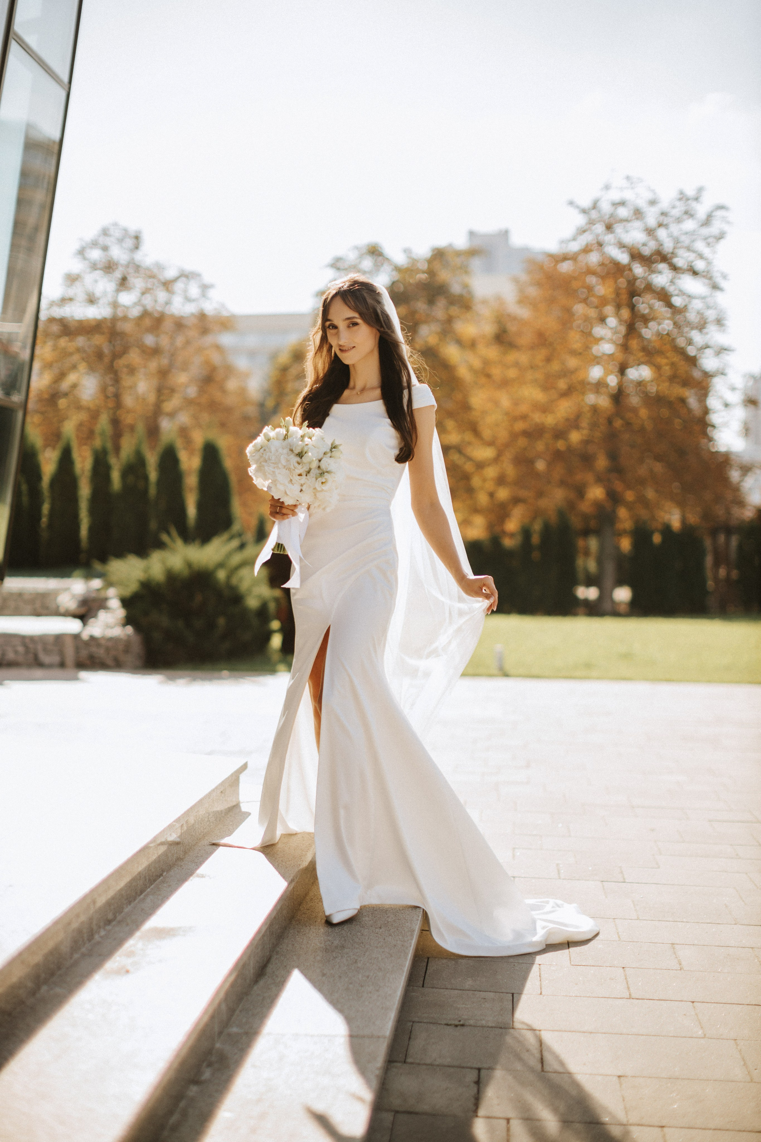 “Una sposa in un elegante abito bianco con uno spacco all'altezza delle cosce scende con grazia i gradini, tenendo in mano un bouquet di fiori bianchi. La luce dorata del sole mette in risalto il tessuto fluente del suo abito, mentre lo sfondo morbido di alberi e vegetazione aggiunge un'atmosfera serena ed elegante”.