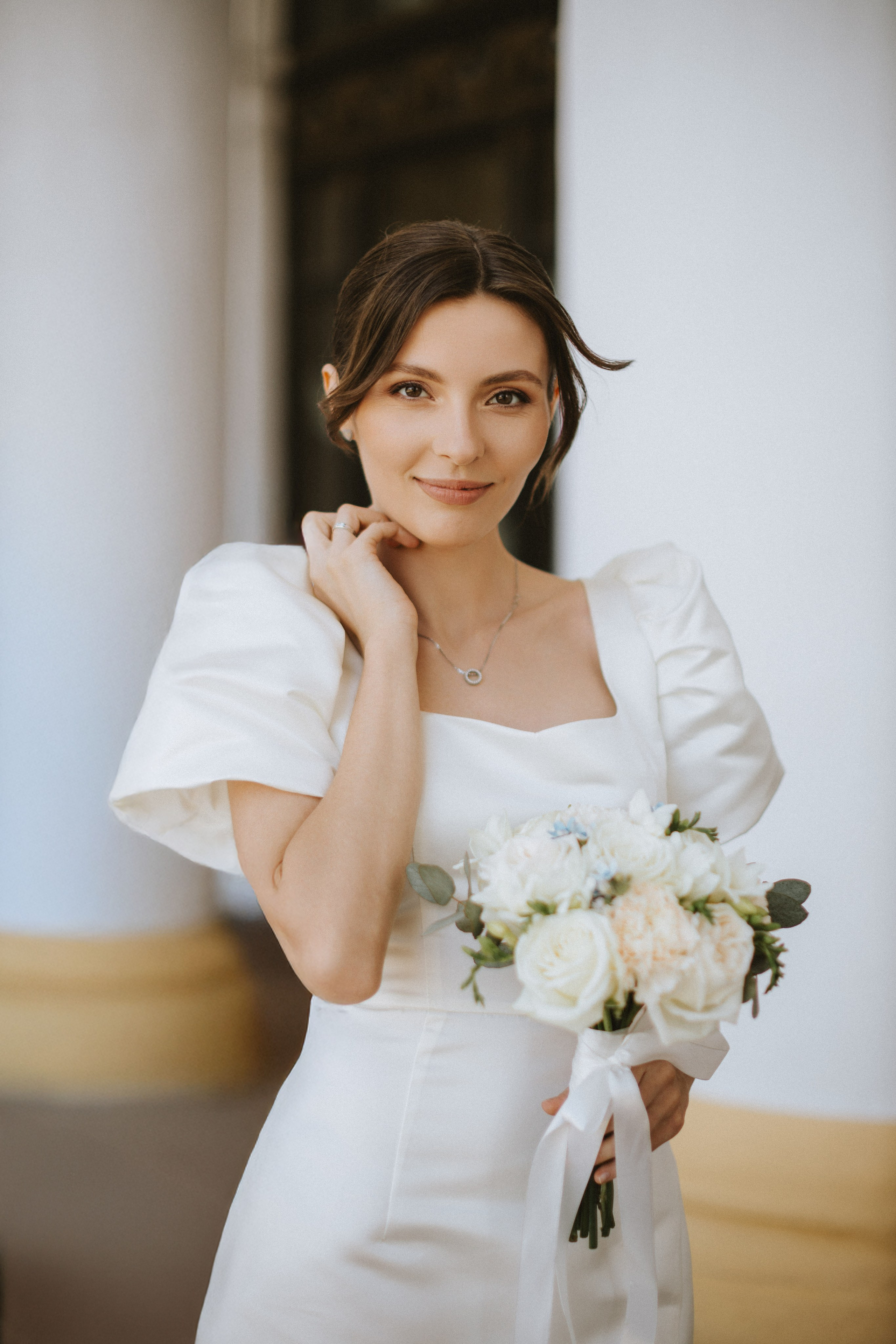 “Una sposa in un abito da sposa chic con maniche a sbuffo, con in mano un bouquet di fiori bianchi e pastello legati con un nastro. Si trova all'aperto con un leggero sorriso, rivolgendo uno sguardo gentile verso la fotocamera. Lo sfondo comprende una parete bianca con un accenno di giallo, che crea un'atmosfera morbida ed elegante”.