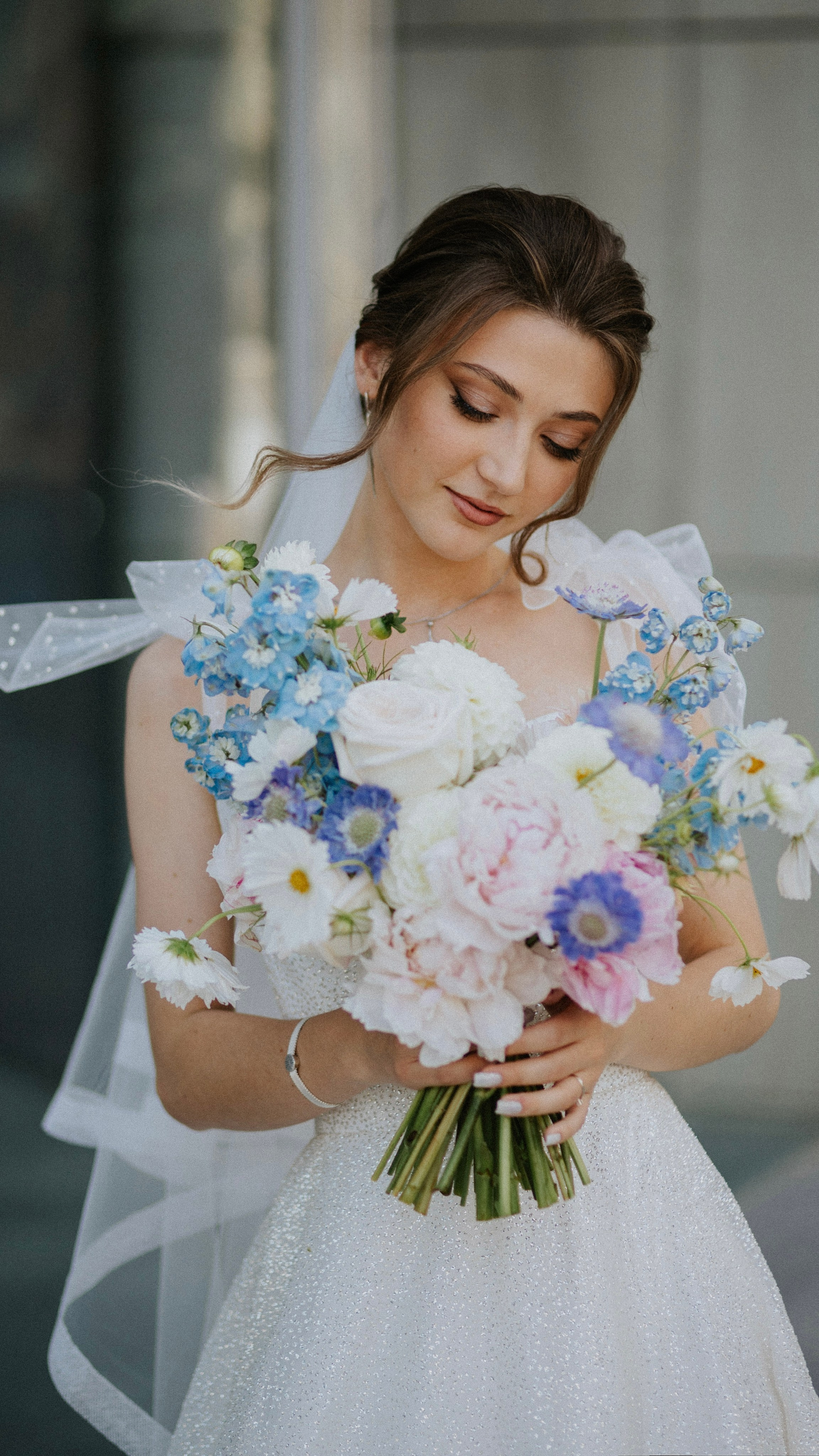 “Una sposa che tiene in mano un bellissimo bouquet di fiori pastello, tra cui peonie, anemoni e margherite, mentre guarda in basso con un tenero sorriso. Indossa un abito da sposa bianco scintillante con un velo delicato, i capelli acconciati a onde sciolte”.