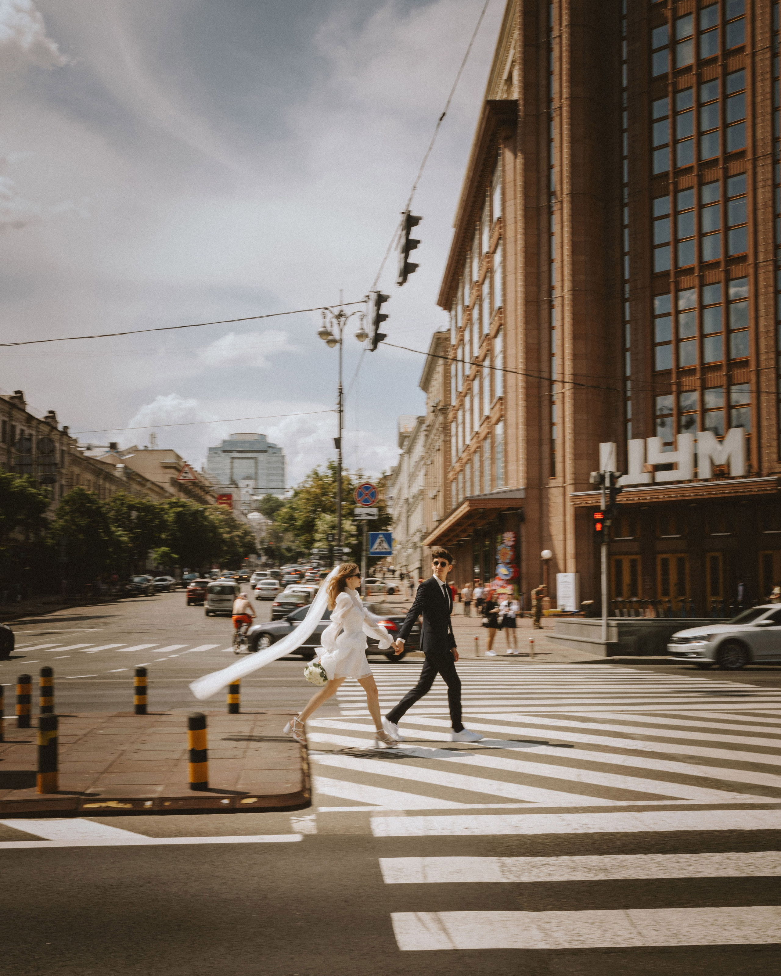 "Modern wedding photoshoot in the city. A stylish bride and groom walk hand in hand across a crosswalk in an urban setting, with wind flowing through the bride’s veil. Candid and dynamic wedding photography."