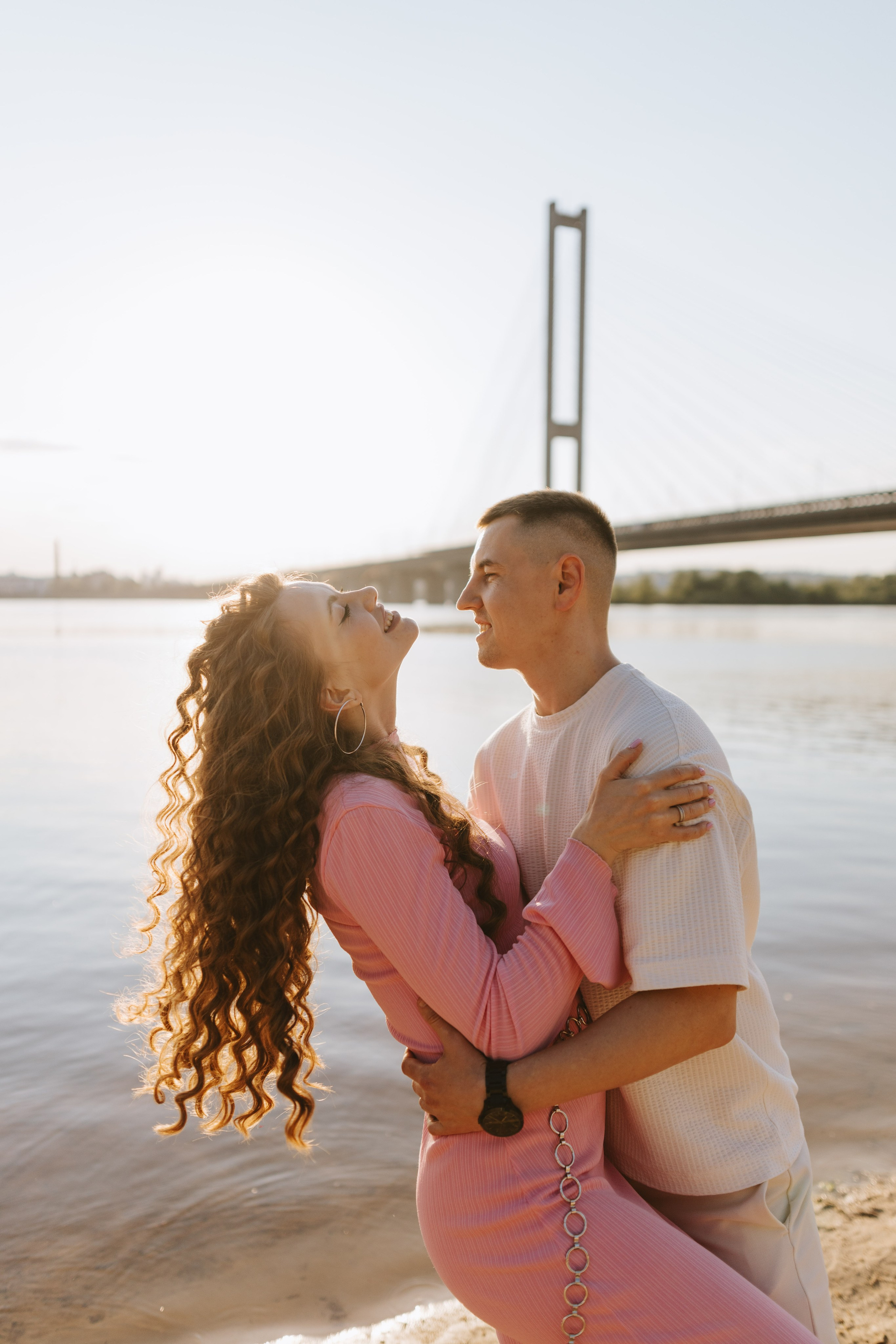 “Una coppia che si gode un momento romantico in riva al mare al tramonto. La donna, vestita con un abito rosa a maniche lunghe con un motivo strutturato, guarda verso il cielo con un'espressione gioiosa, mentre l'uomo, in camicia bianca, la stringe amorevolmente intorno alla vita. Sullo sfondo, un ponte sul fiume e la luce calda e dorata del tramonto illuminano la scena, creando un'atmosfera serena e intima”.