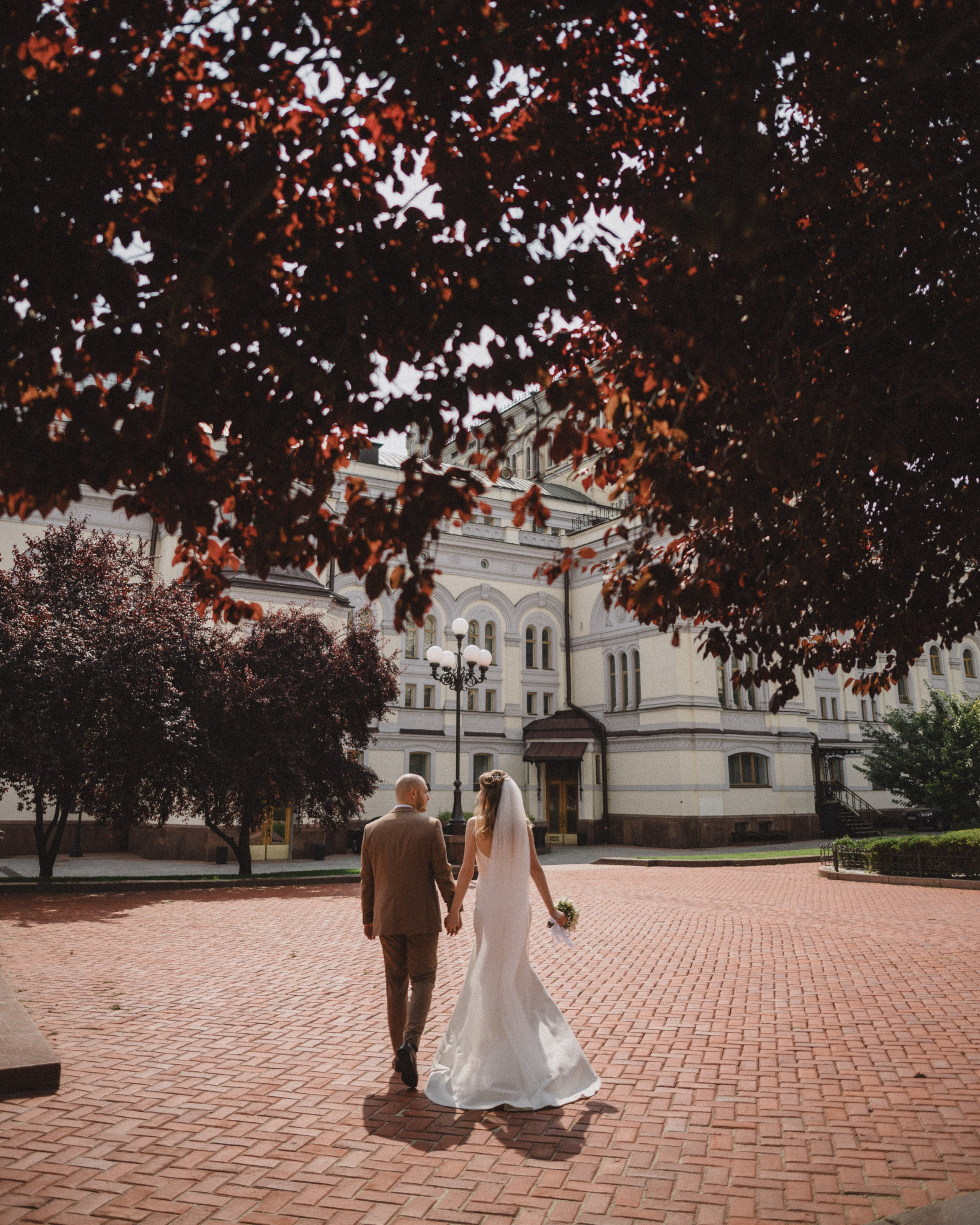 The bride and groom walk down the path holding hands, with a historic building and trees with red leaves in the background.”