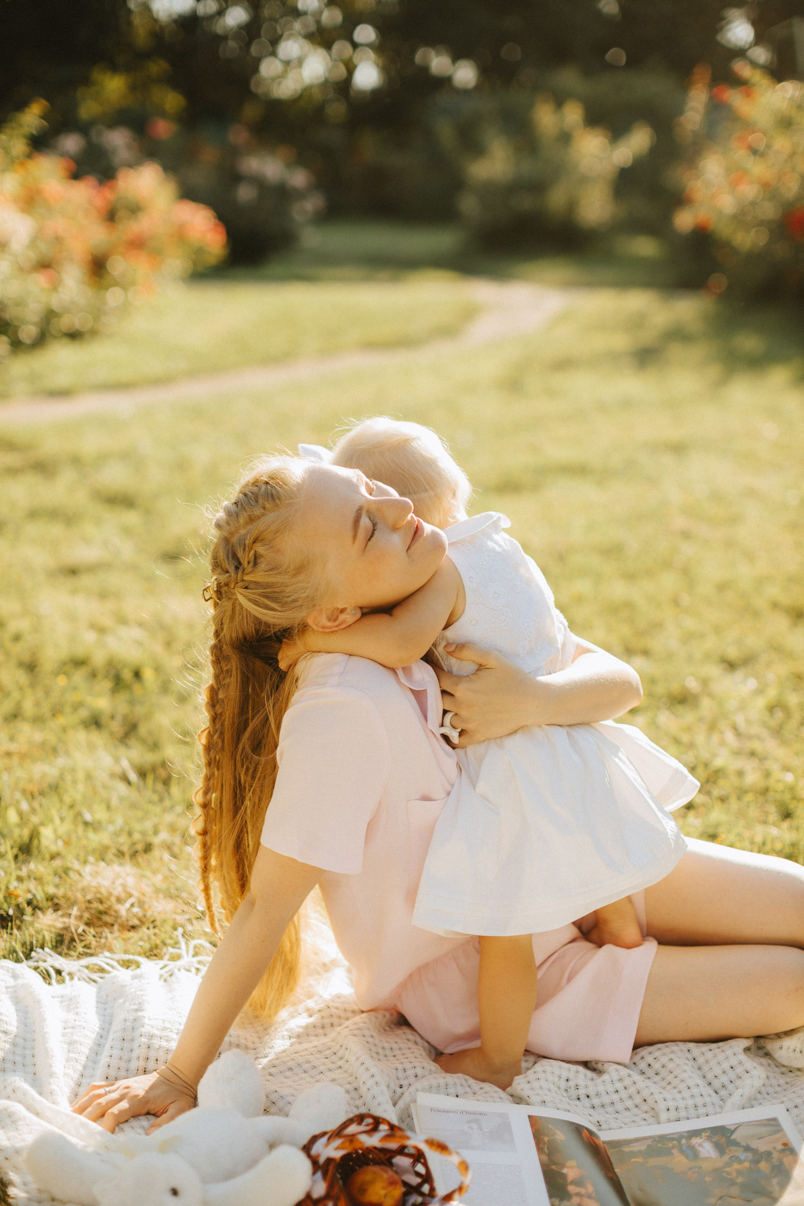 “Una madre che abbraccia il suo bambino all'aperto, seduta su una coperta in un giardino con la luce del sole che filtra, mostrando un tenero momento di affetto”.
