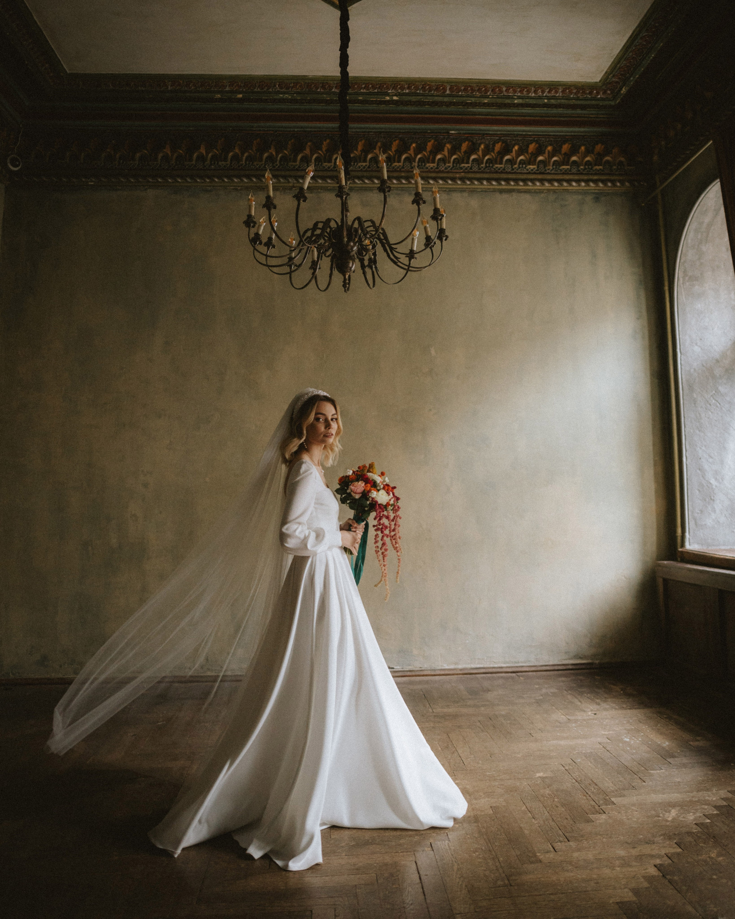 “The bride in a long dress and veil, holding a bouquet, walks through the interior of an old room.”