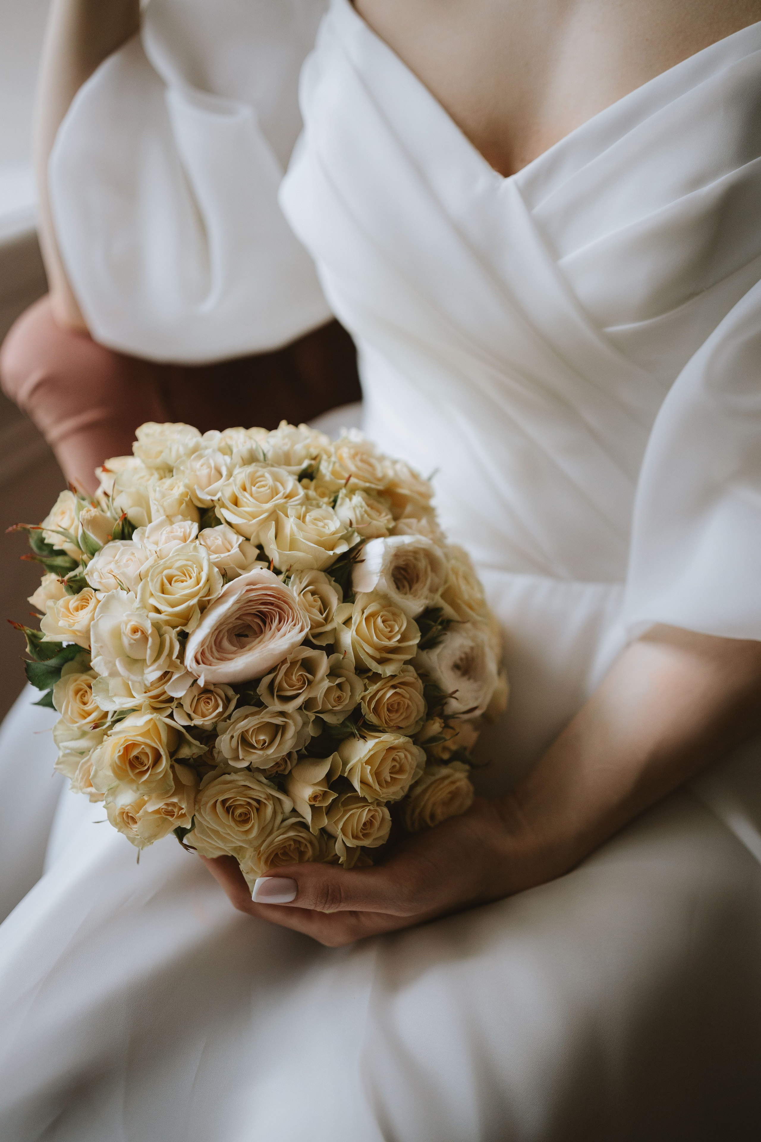 “Primo piano di una sposa che tiene in mano un bellissimo bouquet di rose morbide color crema. Le rose sono delicatamente raggruppate, con alcuni fiori chiari che spiccano tra gli altri. Sullo sfondo si intravede l'abito da sposa in raso bianco della sposa, dal tessuto morbido e fluente. L'elegante semplicità del bouquet contrasta magnificamente con la texture lussuosa dell'abito”.