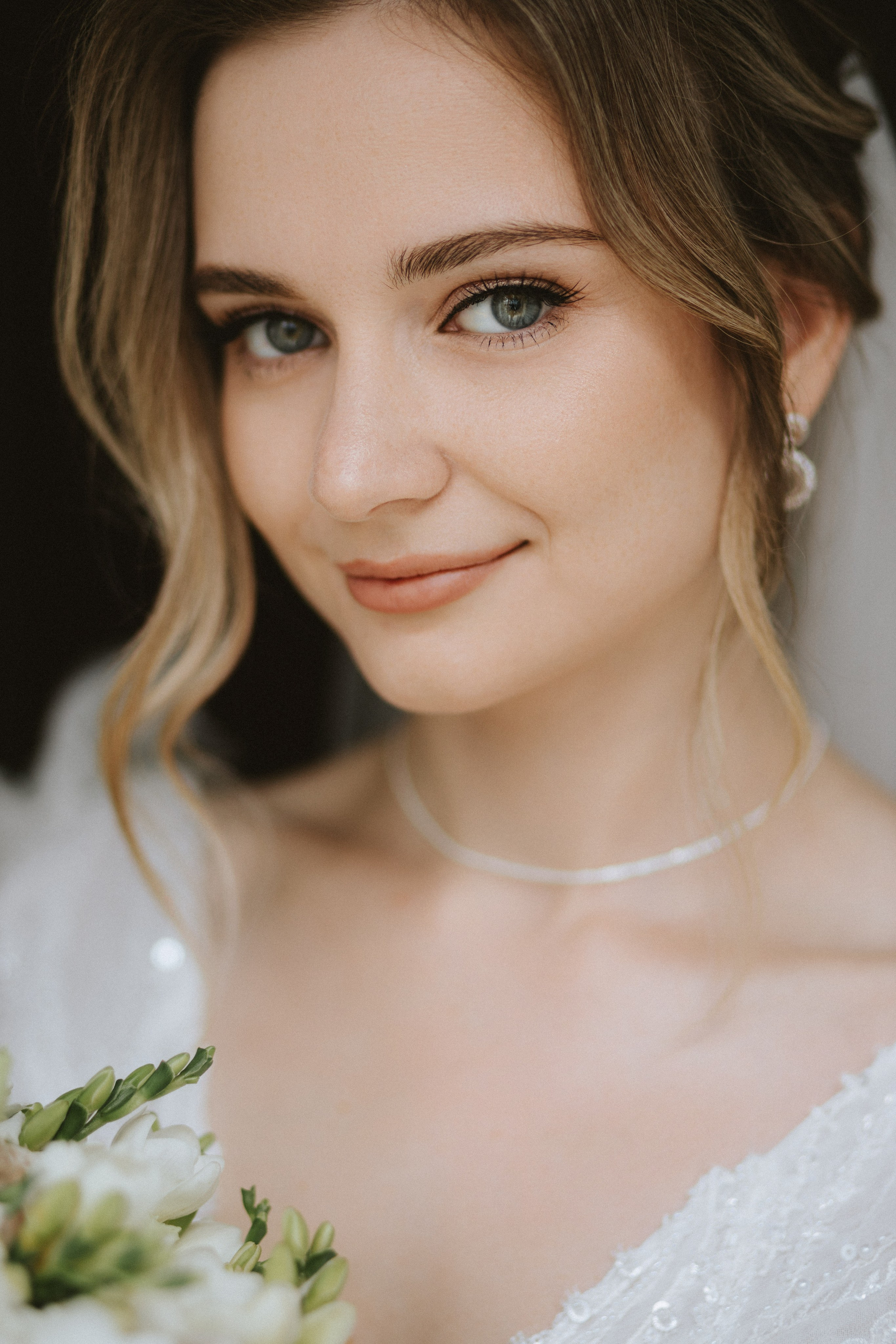 “Primo piano di una bellissima sposa con scintillanti occhi azzurri e morbidi riccioli, che indossa una delicata collana e orecchini. La sposa tiene in mano un bouquet di fiori bianchi e verdi, perfetto per un matrimonio romantico in Toscana, Italia”.