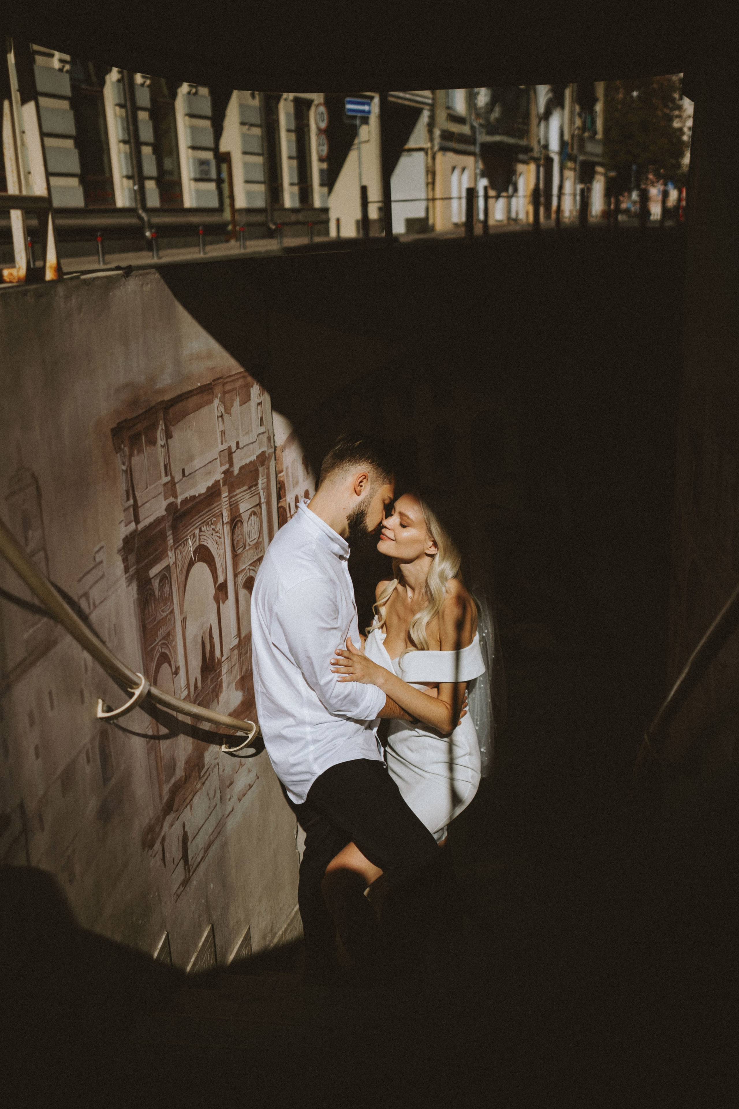 “Un momento romantico tra una coppia in piedi in una tromba delle scale, immersa in una tenue luce solare. La sposa, in abito bianco off-the-shoulder e velo, condivide un tenero bacio con lo sposo, vestito con una camicia bianca. Un murale di architettura antica decora la parete alle loro spalle, creando un'atmosfera intima e senza tempo”.