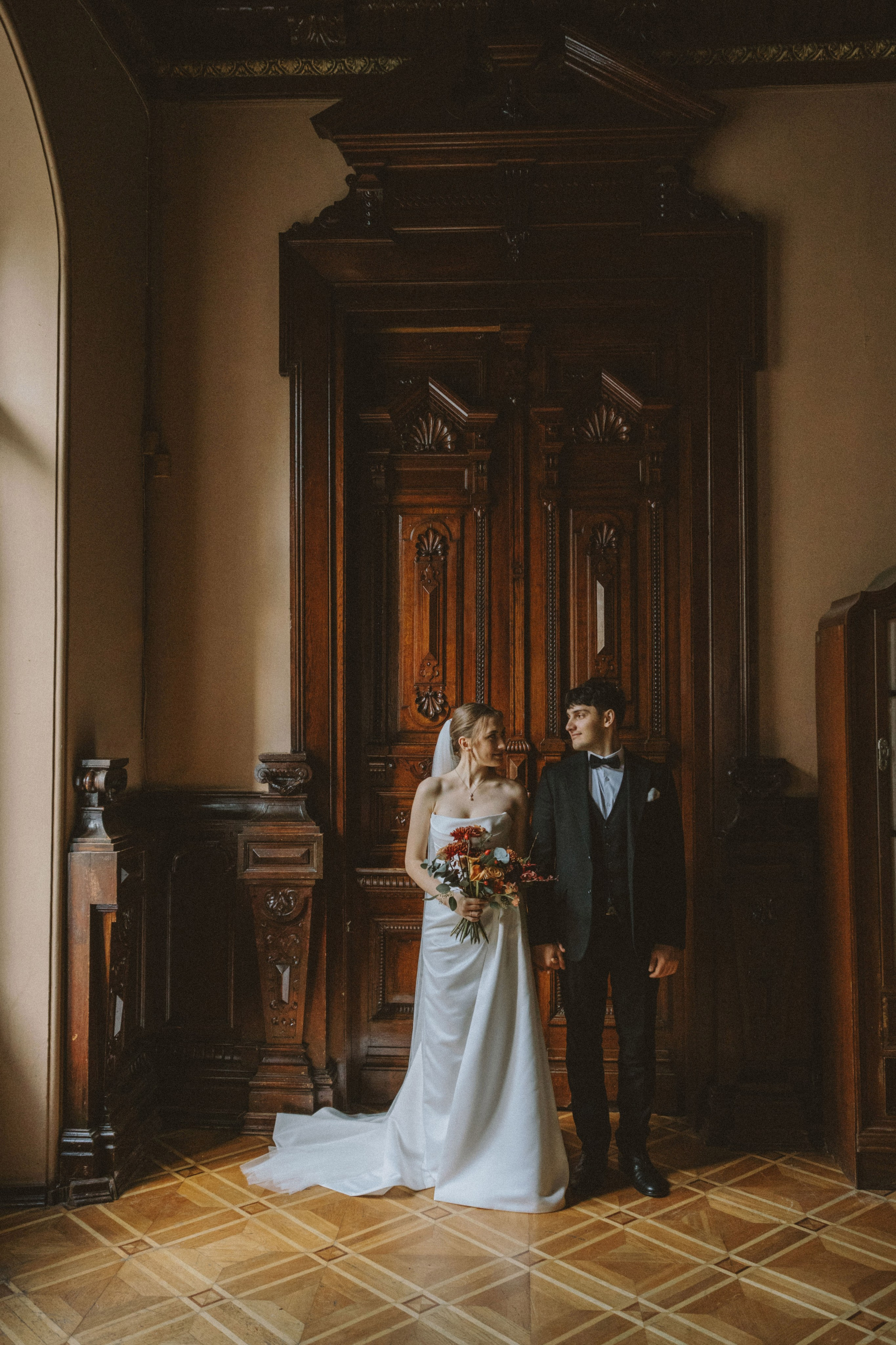 “Una sposa e uno sposo stanno insieme davanti a un armadio vintage in legno e si guardano sorridenti. La sposa tiene in mano un bouquet di fiori e lo sposo è vestito con un abito nero e un papillon. La stanza ha un arredamento elegante con dettagli in legno e luci soffuse”.