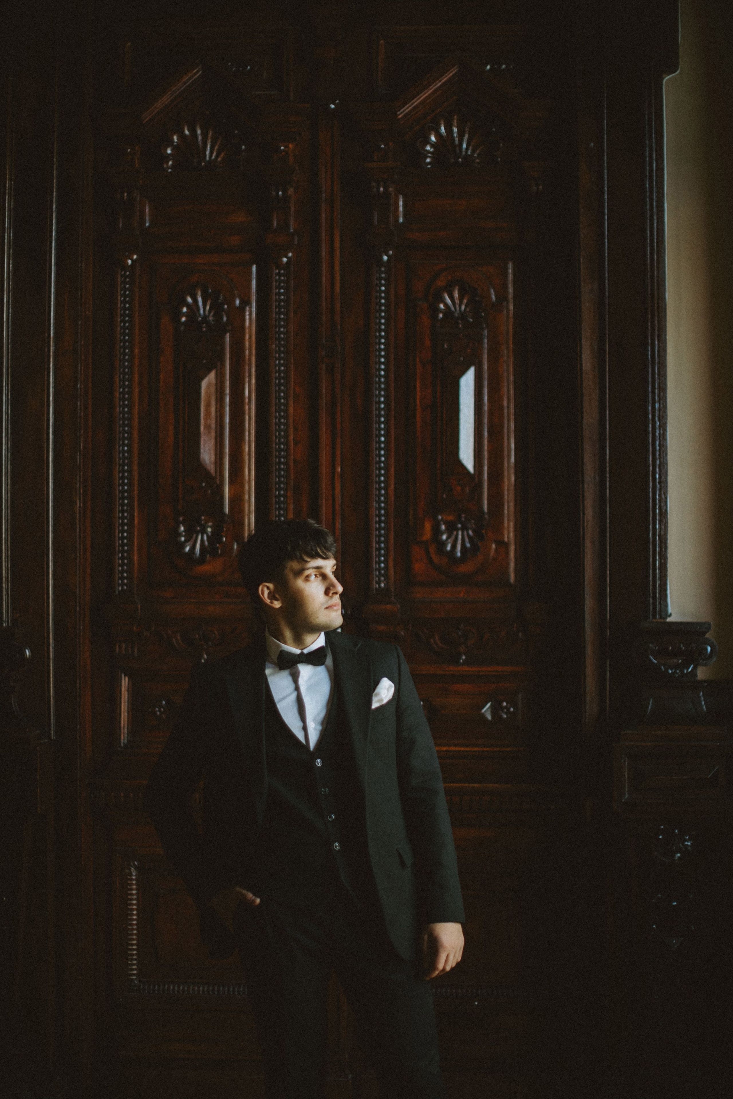 A groom stands confidently in front of an intricately carved wooden door, dressed in a black tuxedo with a white shirt and bow tie. His gaze is directed to the side, creating a contemplative and elegant mood.”