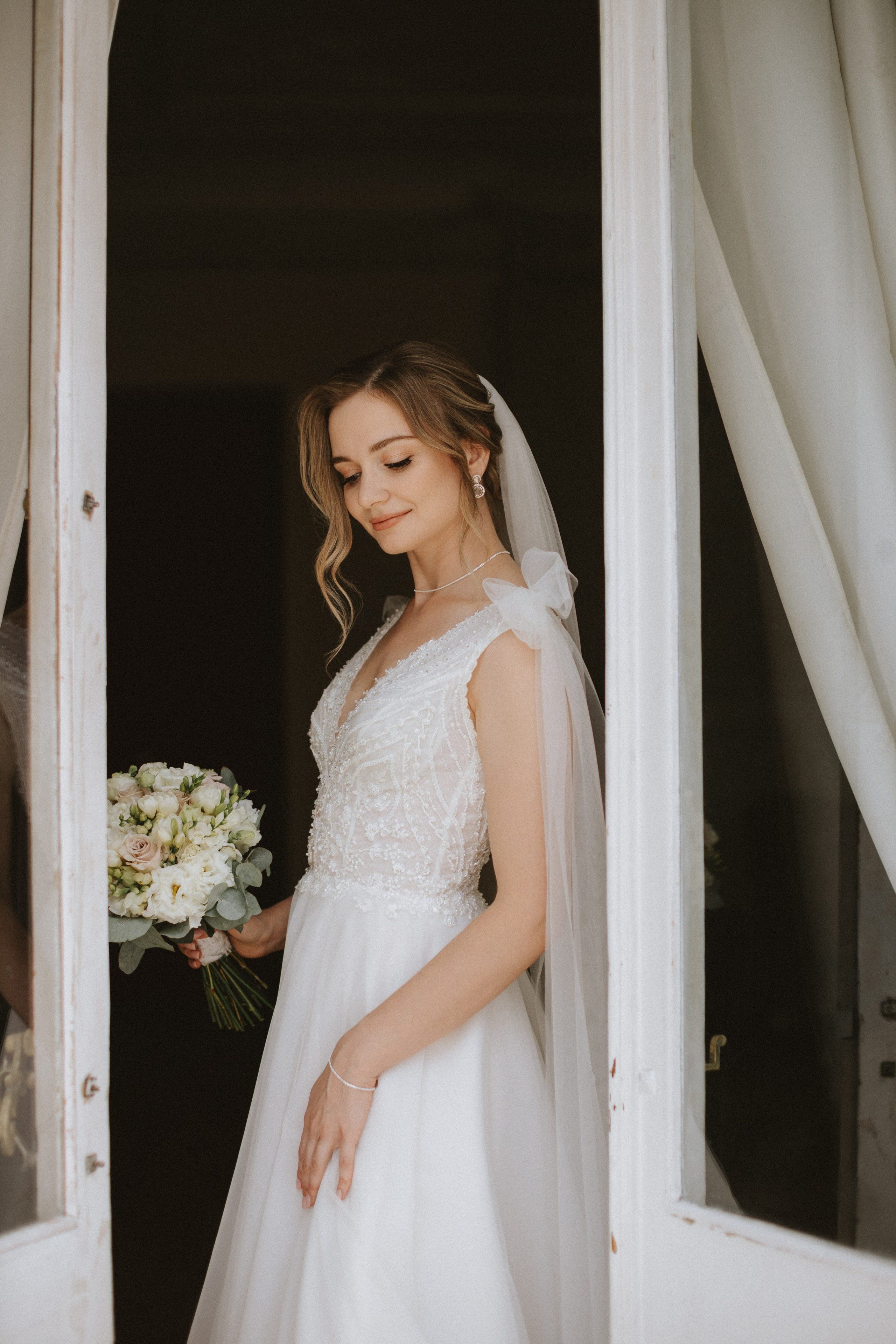 “Una sposa si affaccia con grazia a una finestra, tenendo in mano un bouquet di fiori bianchi e rosa pallido, con la luce del sole che illumina i suoi lineamenti delicati. Indossa un abito elegante ornato da intricate perline e un velo fluente con un fiocco, la testa leggermente inclinata mentre sorride dolcemente”.