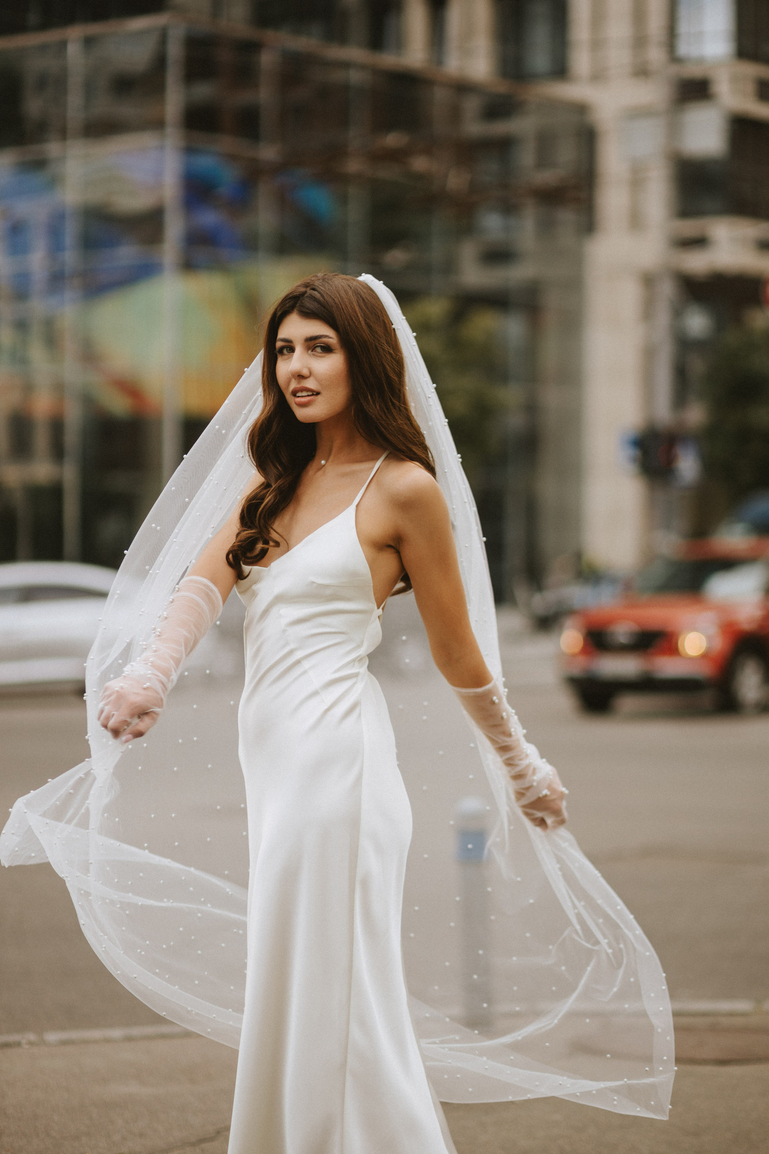 “Una sposa in un elegante abito bianco satinato si trova all'aperto, con un velo impreziosito da perle che le scorre dietro. Indossa guanti trasparenti e guarda direttamente verso l'obiettivo, con i capelli acconciati in morbide onde. Lo sfondo urbano e il traffico si confondono delicatamente, mettendo in risalto la sua graziosa presenza contro il moderno ambiente cittadino”.