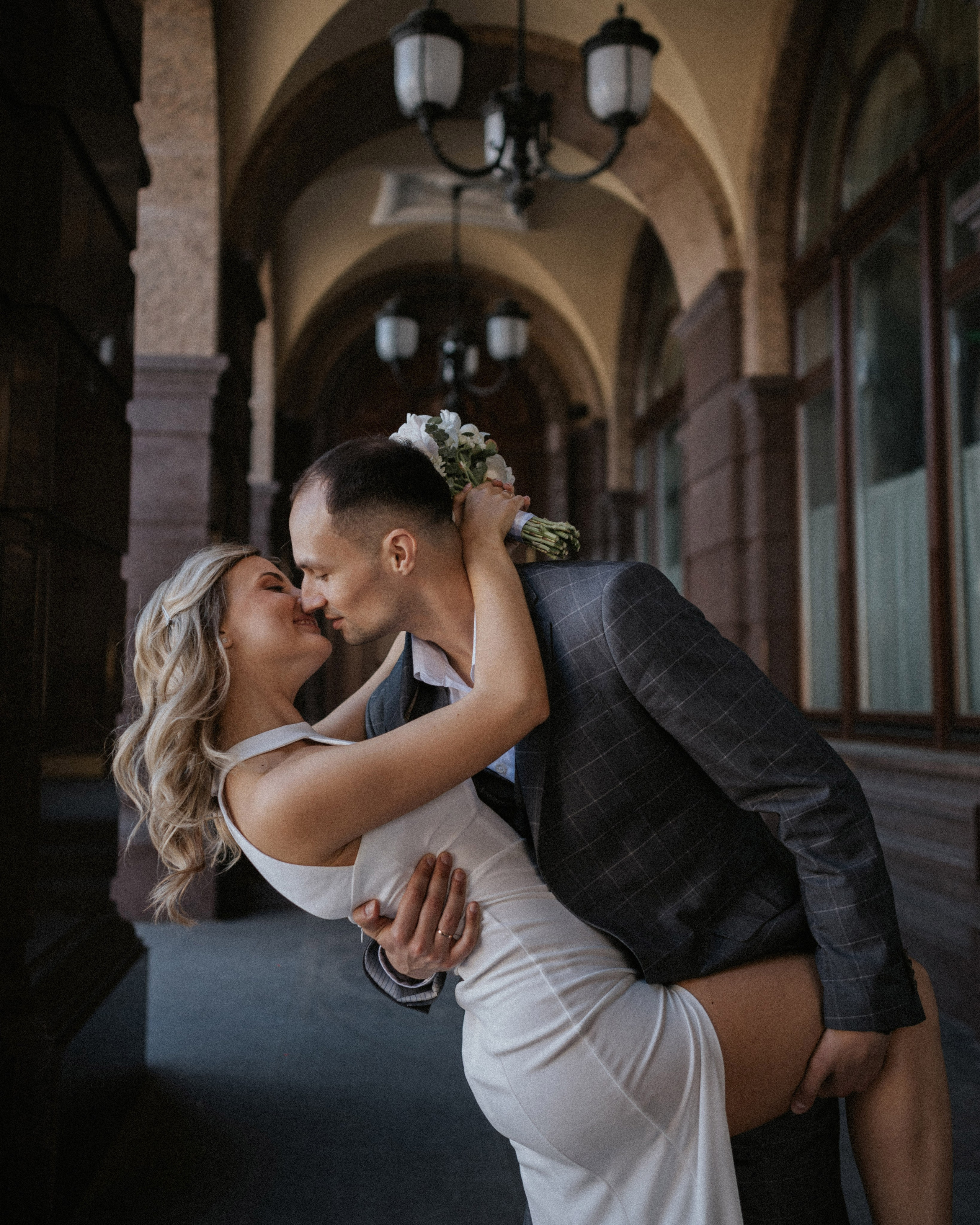 “Una sposa e uno sposo che si scambiano un bacio romantico in un arco all'aperto. La sposa, vestita con un abito da sposa bianco, viene sollevata dallo sposo, che indossa un abito grigio a scacchi. L'atmosfera è calda e intima, con luci soffuse provenienti da lampade a sospensione sullo sfondo”.