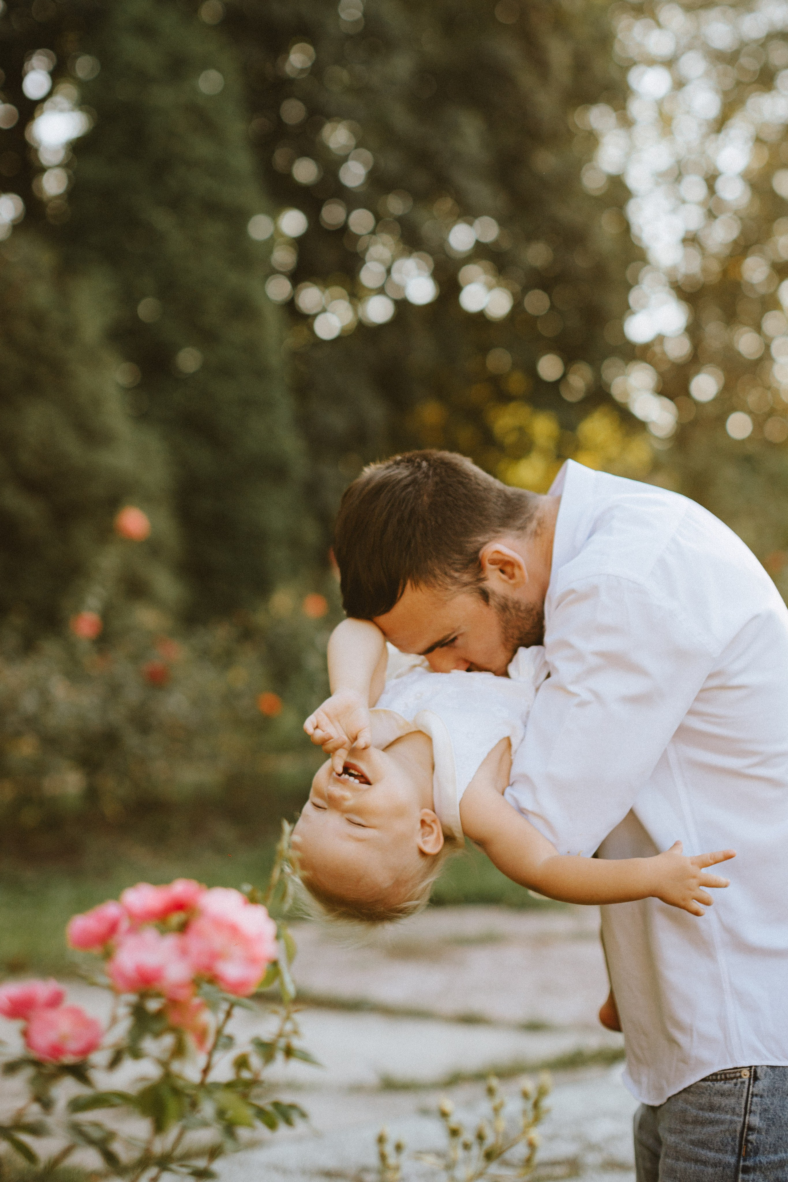 Un padre gioioso che solleva la sua ridente figlioletta a testa in giù, con la bambina che raggiunge giocosamente il viso del padre. Lo sfondo è caratterizzato da una vegetazione morbida e sfocata e da fiori che sbocciano, catturando un momento di amore e divertimento nella natura”.