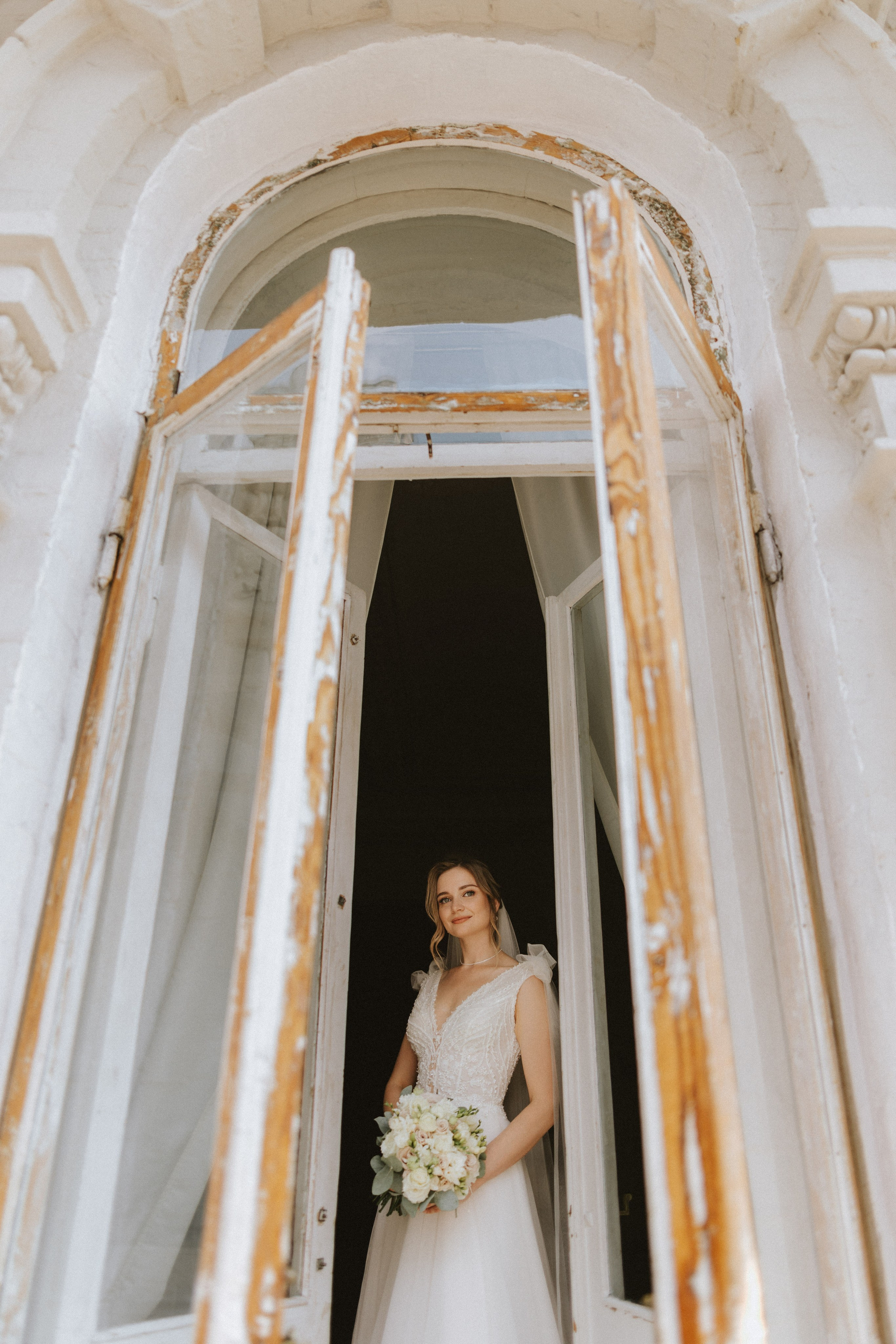 “La sposa si affaccia con grazia alla finestra aperta, tenendo in mano il suo bouquet mentre sorride serenamente. Il suo delicato abito da sposa e il suo morbido velo creano un bellissimo momento di eleganza, incorniciato dall'affascinante architettura, perfetto per un matrimonio in una location storica.”