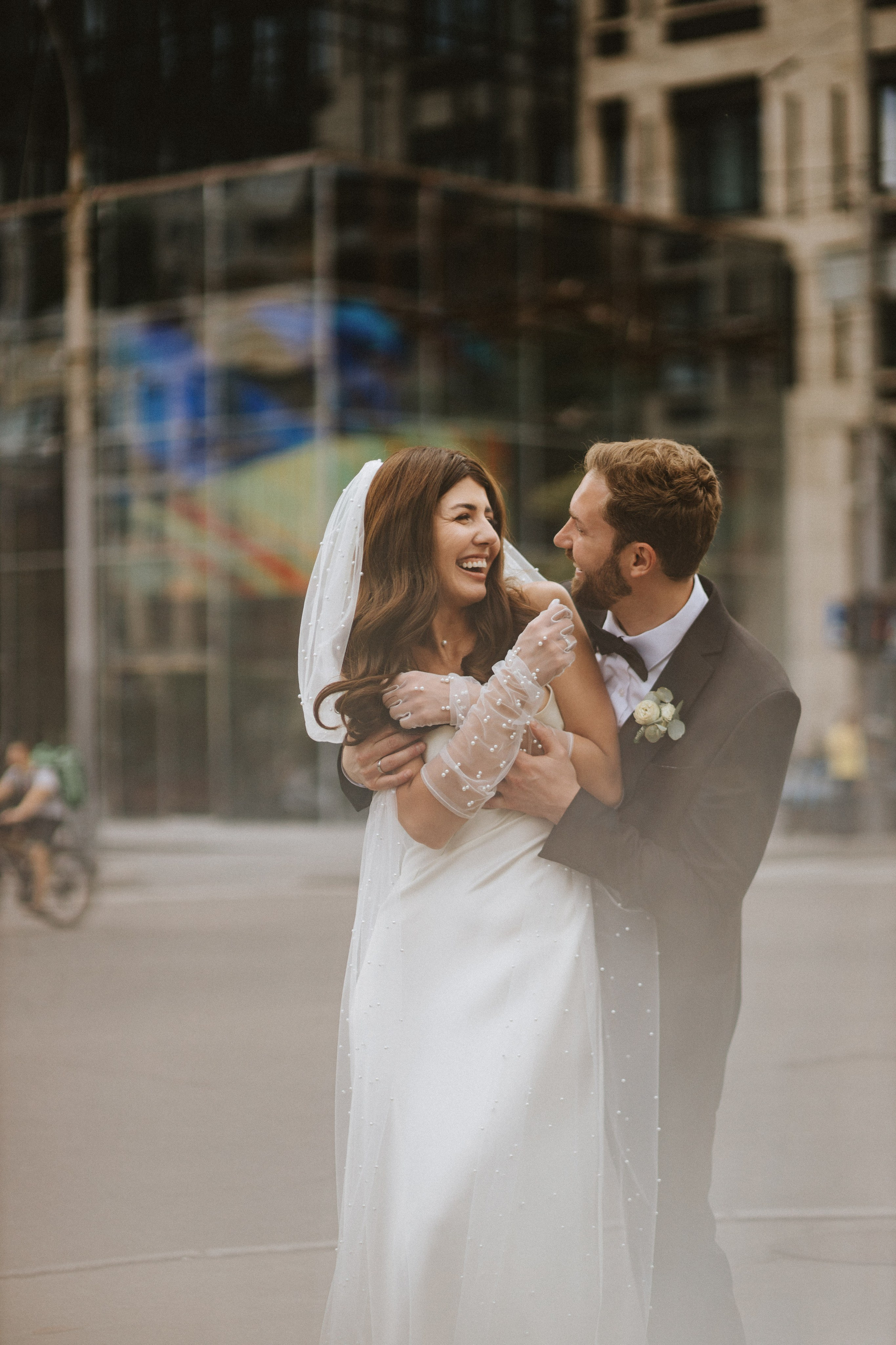 “Un momento gioioso catturato mentre gli sposi ridono insieme nel mezzo della città, con il velo della sposa e il suo abito di perline che aggiungono un tocco di eleganza. La felicità della coppia è splendidamente evidenziata dallo sfondo urbano, rendendo questo scatto perfetto per un matrimonio moderno”.