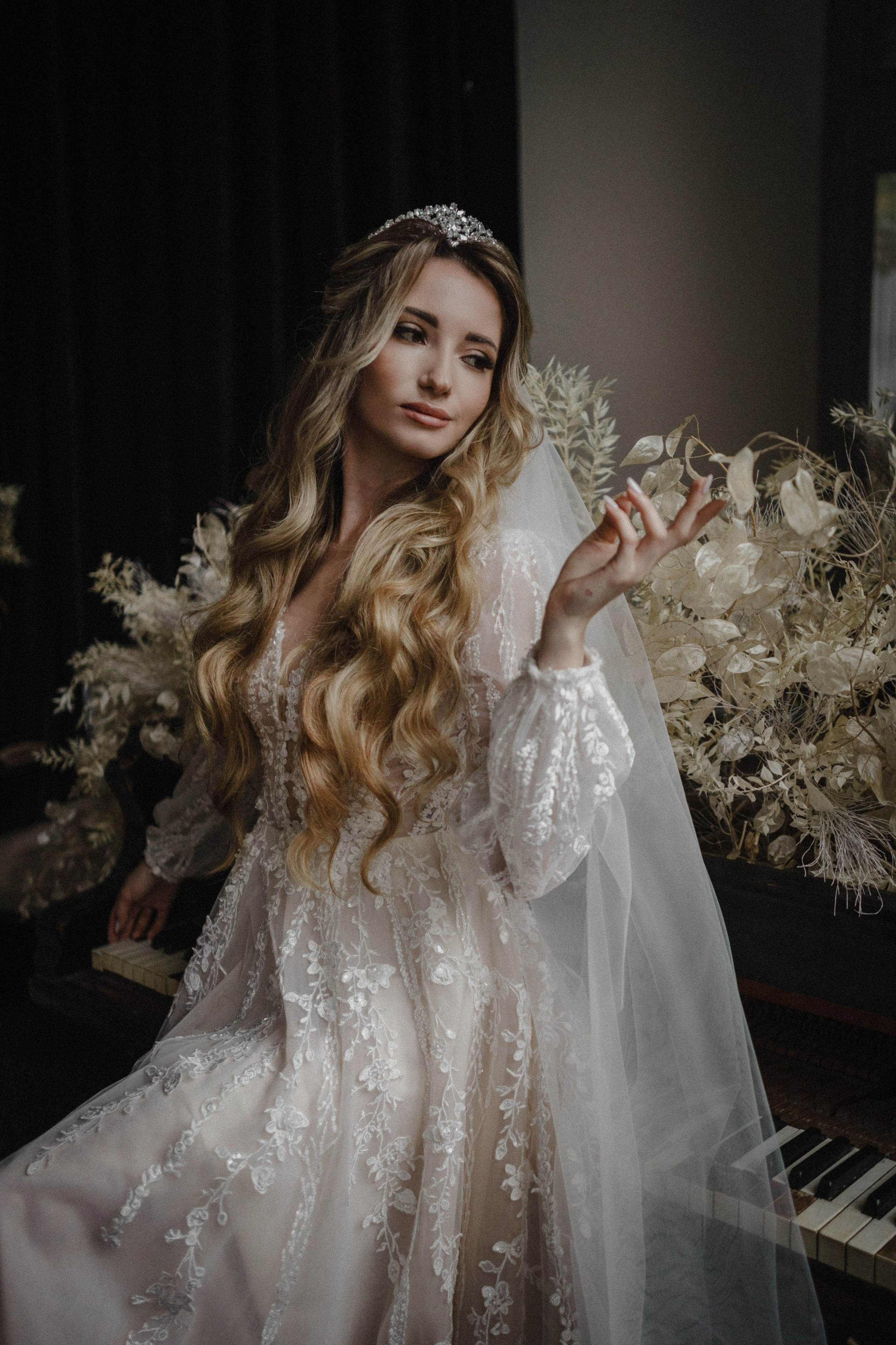 “Una sposa in un delicato abito da sposa di pizzo con intricati ricami floreali siede accanto a un pianoforte d'epoca. Guarda pensierosa in lontananza, con la mano sollevata delicatamente, tenendo un ramo di verde tenero. I suoi lunghi e fluenti capelli sono ornati da un diadema scintillante e il velo le ricade con grazia sulle spalle. L'atmosfera è serena e romantica, con luci soffuse che esaltano la scena elegante”.