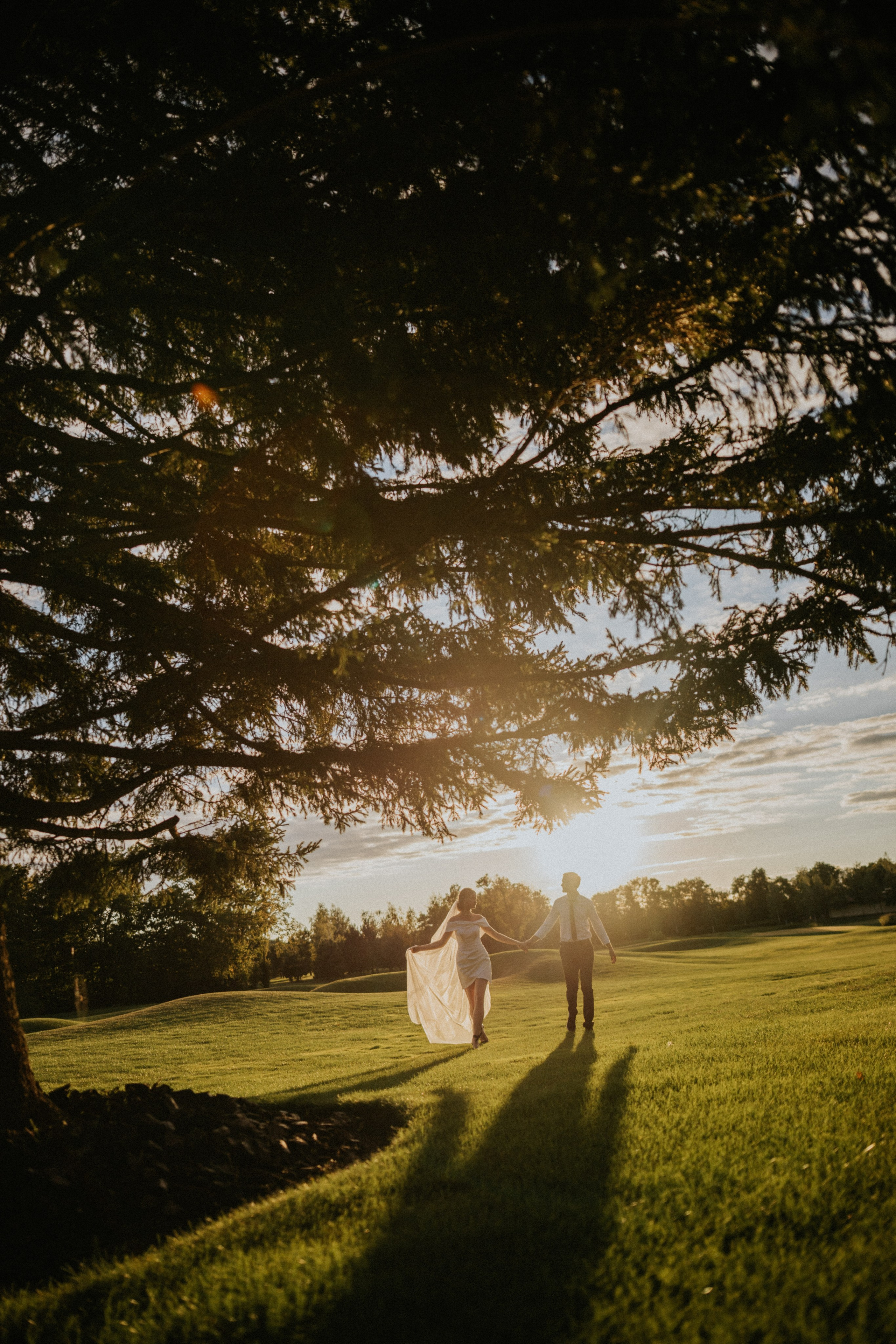 “Una coppia cammina mano nella mano attraverso un campo verde e lussureggiante, immerso in una calda luce dorata durante il tramonto. L'abito fluente della sposa cattura la brezza, creando un momento magico sotto gli alberi, perfetto per un matrimonio romantico in Italia.”