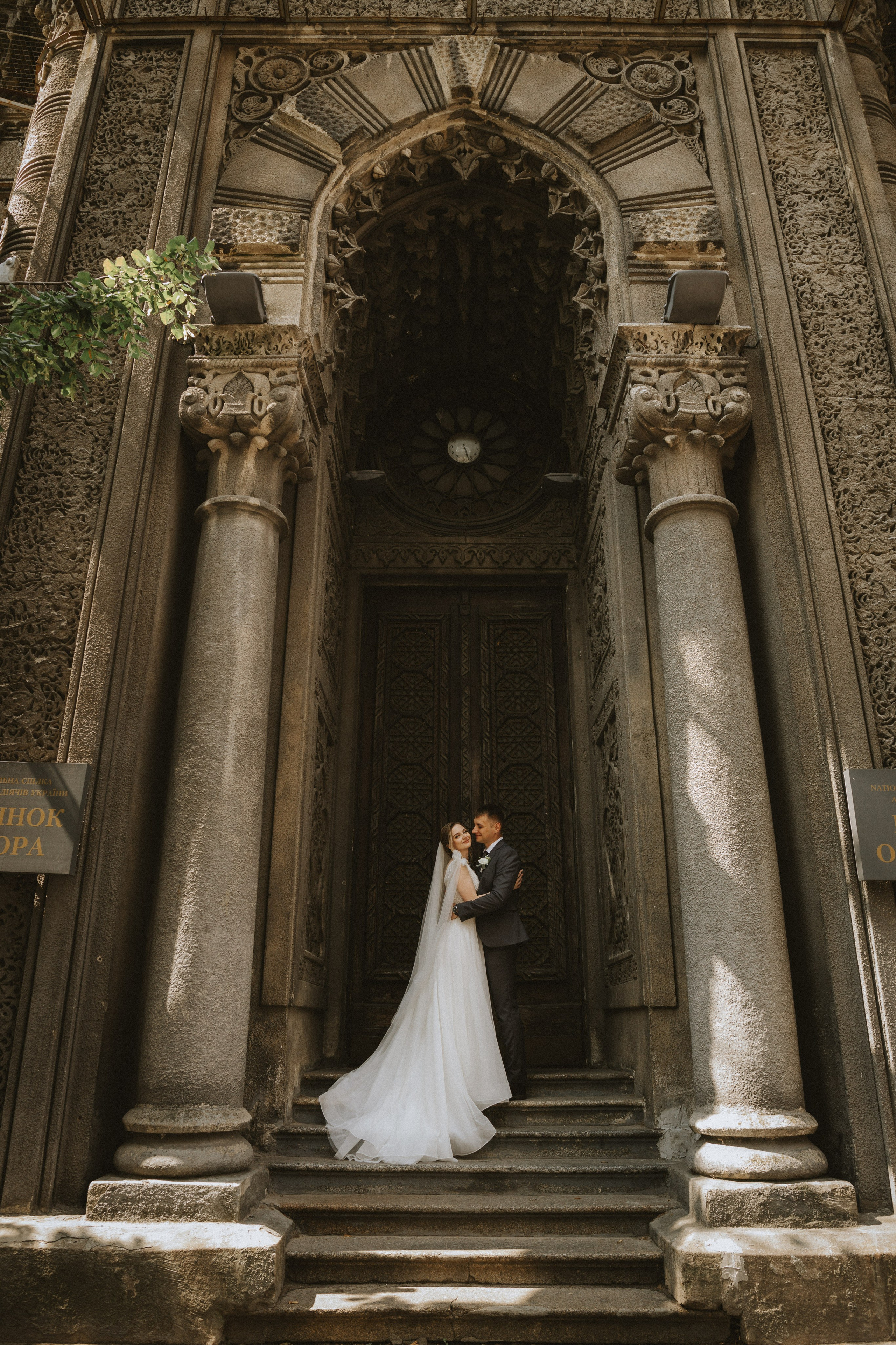 “Una coppia si trova sui gradini di un edificio splendidamente decorato e condivide un momento di intimità. La sposa, vestita con un elegante abito bianco e un lungo velo, e lo sposo, in abito elegante, si sorridono sotto il grande arco. L'architettura dettagliata e la luce soffusa del sole creano uno sfondo romantico e senza tempo”.