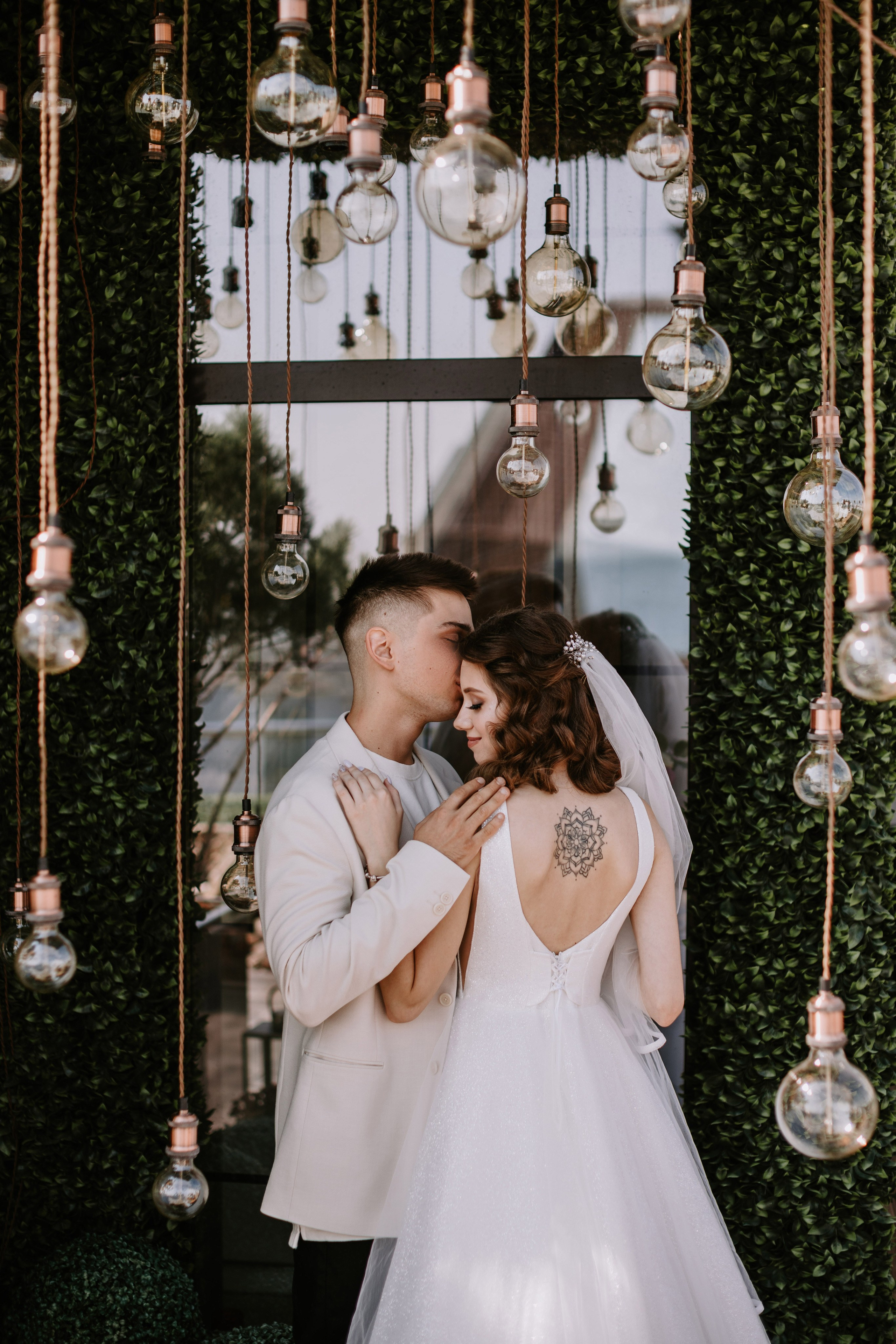 “Un momento di tenerezza quando lo sposo bacia la fronte della sposa, in piedi insieme davanti a un bellissimo sfondo con lampadine sospese e verde. L'abito elegante della sposa e l'intricato tatuaggio sulla schiena aggiungono un tocco unico a questo intimo scatto di matrimonio”.