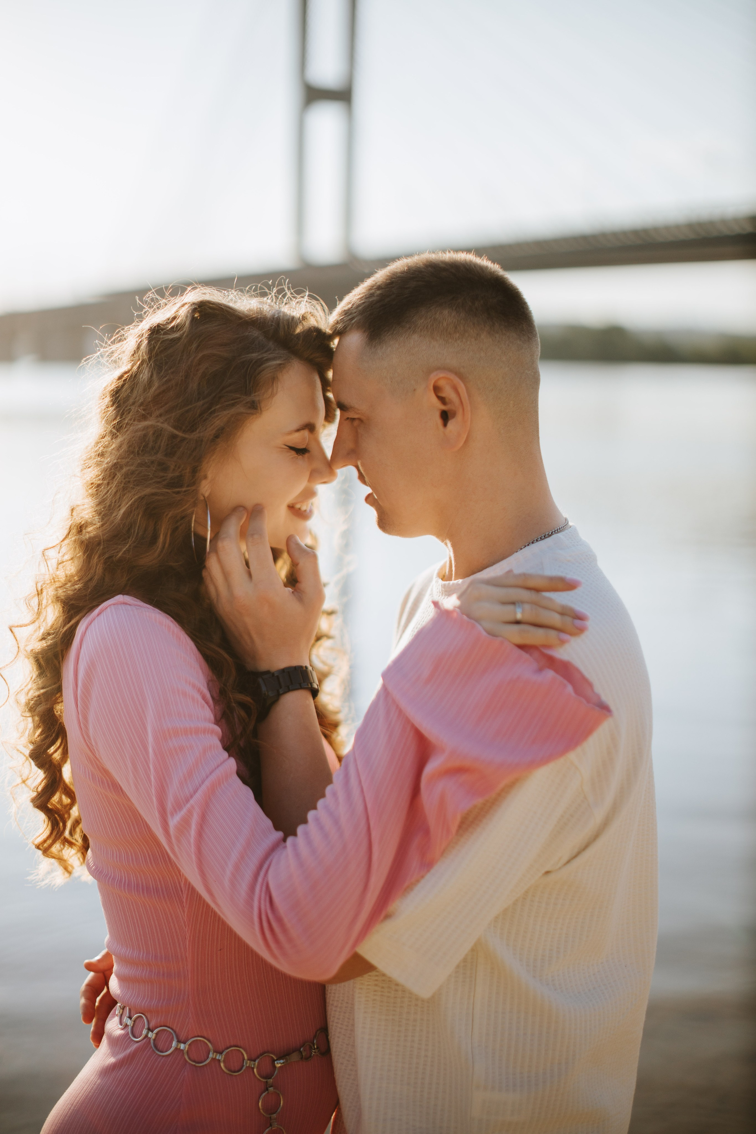 Una coppia in un romantico abbraccio, che condivide un momento di tenerezza al tramonto in riva al fiume, con il ponte sullo sfondo. Sorridono, con le fronti che si toccano e gli occhi chiusi”.