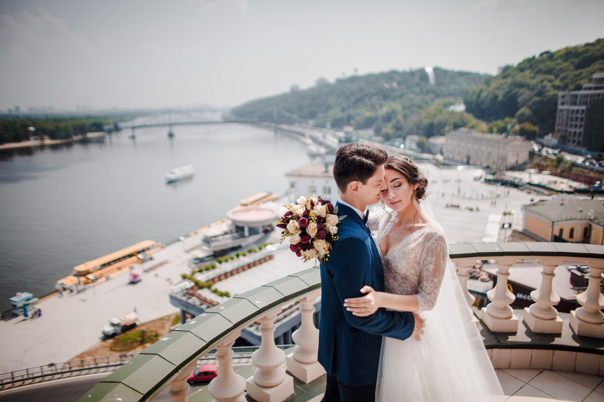 “Una bellissima foto di matrimonio di una coppia che si abbraccia sullo splendido sfondo del fiume e dello skyline della città. La sposa indossa un delicato abito da sposa in pizzo e tiene in mano un bouquet di rose rosse, bianche e crema, mentre lo sposo è vestito con un abito blu. Si trovano su un balcone, crogiolandosi nella luce del sole, con il fiume e gli edifici sullo sfondo, catturando un momento di amore e intimità”.