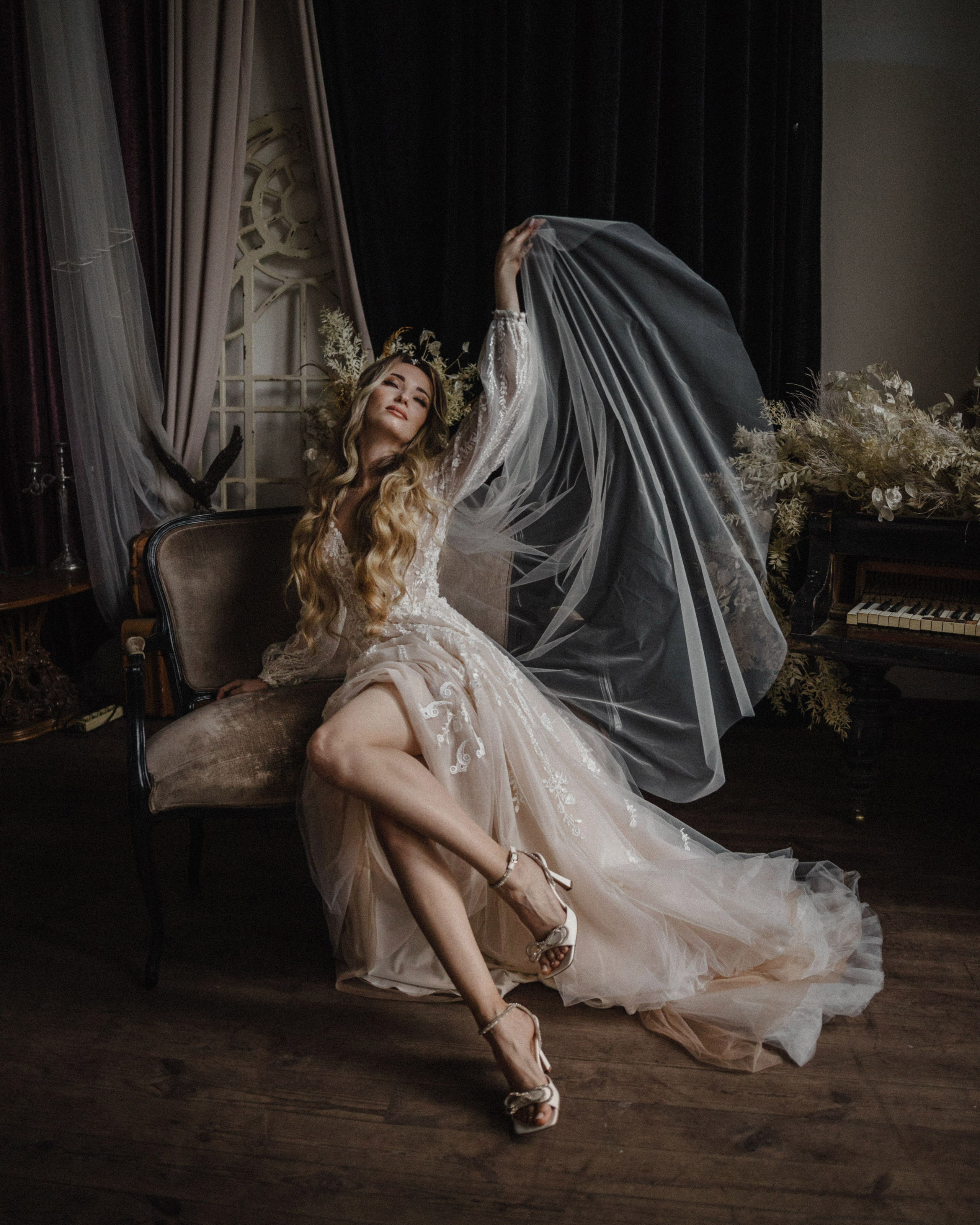 "A bride in an elegant wedding dress with a long train sits on a vintage chair, holding her veil above her head, in a romantic setting with flowers and a piano in the background."