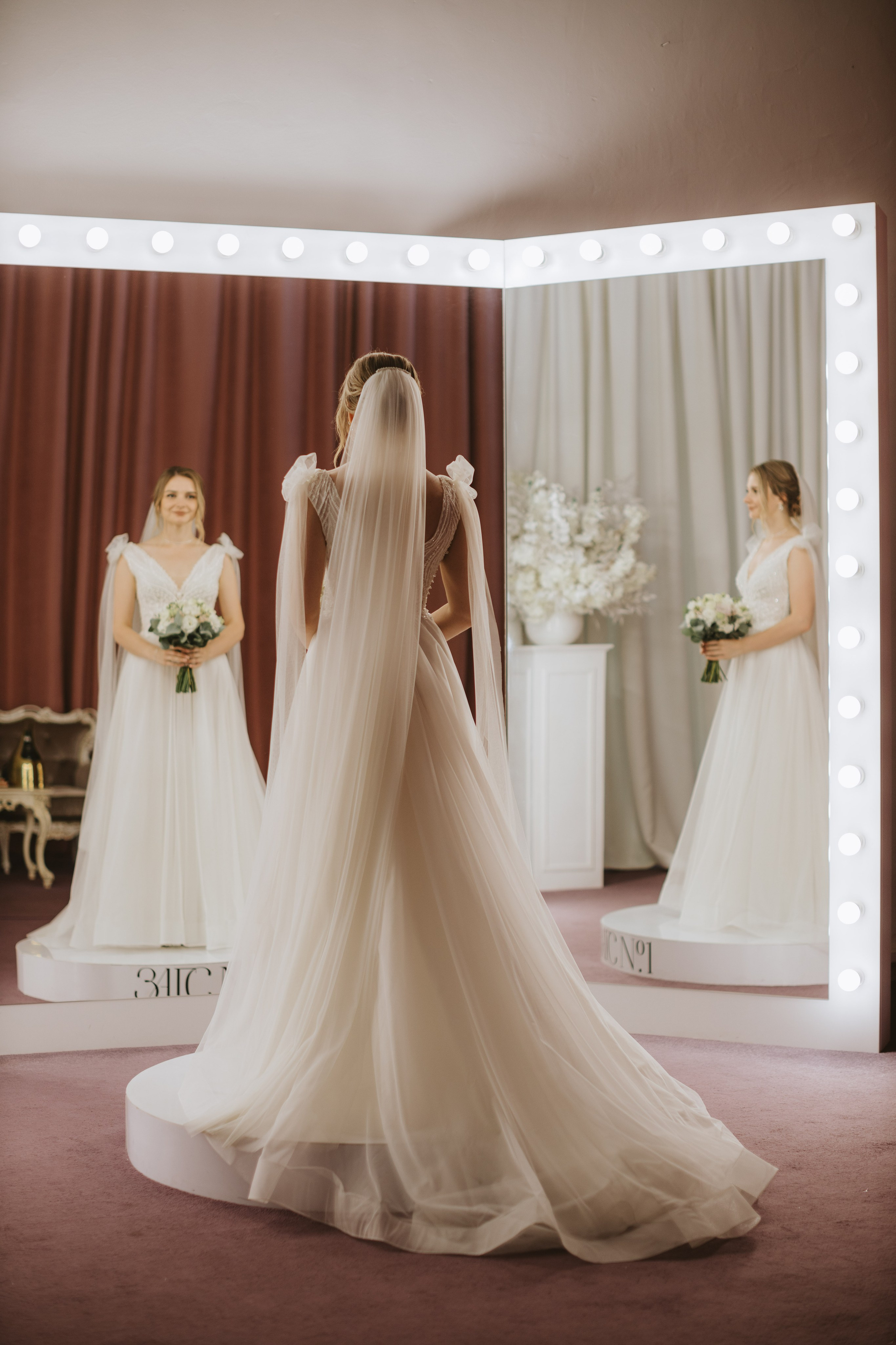 “Una sposa in piedi davanti a uno specchio, che ammira il suo riflesso. Indossa un fluente abito da sposa bianco con delicati fiocchi sulle spalle e un lungo velo. L'illuminazione delle lampadine dello specchio mette in risalto le linee aggraziate dell'abito, mentre tiene in mano un piccolo bouquet di fiori”.