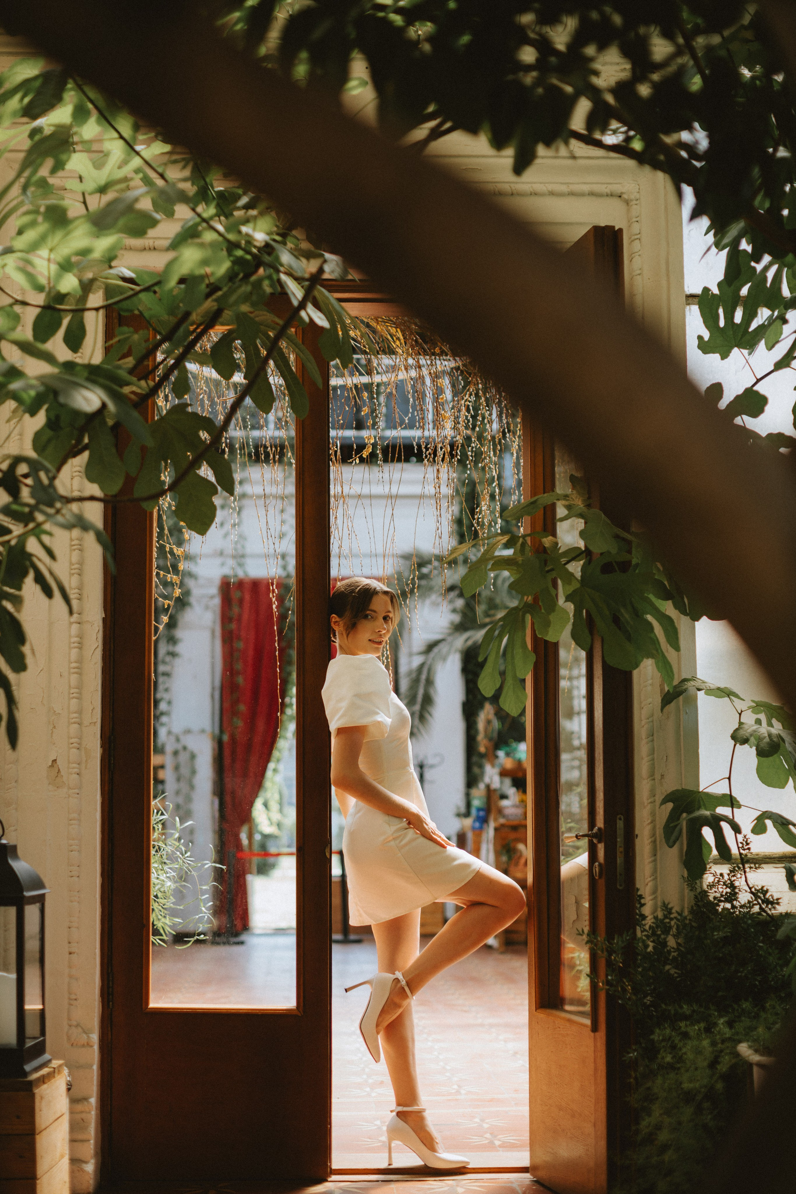 “Una sposa in abito bianco minimalista e tacchi assume una posa aggraziata in un ingresso, incorniciata da una vegetazione lussureggiante. La luce del sole filtra attraverso le piante, creando un'atmosfera morbida e romantica, mentre lei si guarda alle spalle con un sorriso gentile”.
