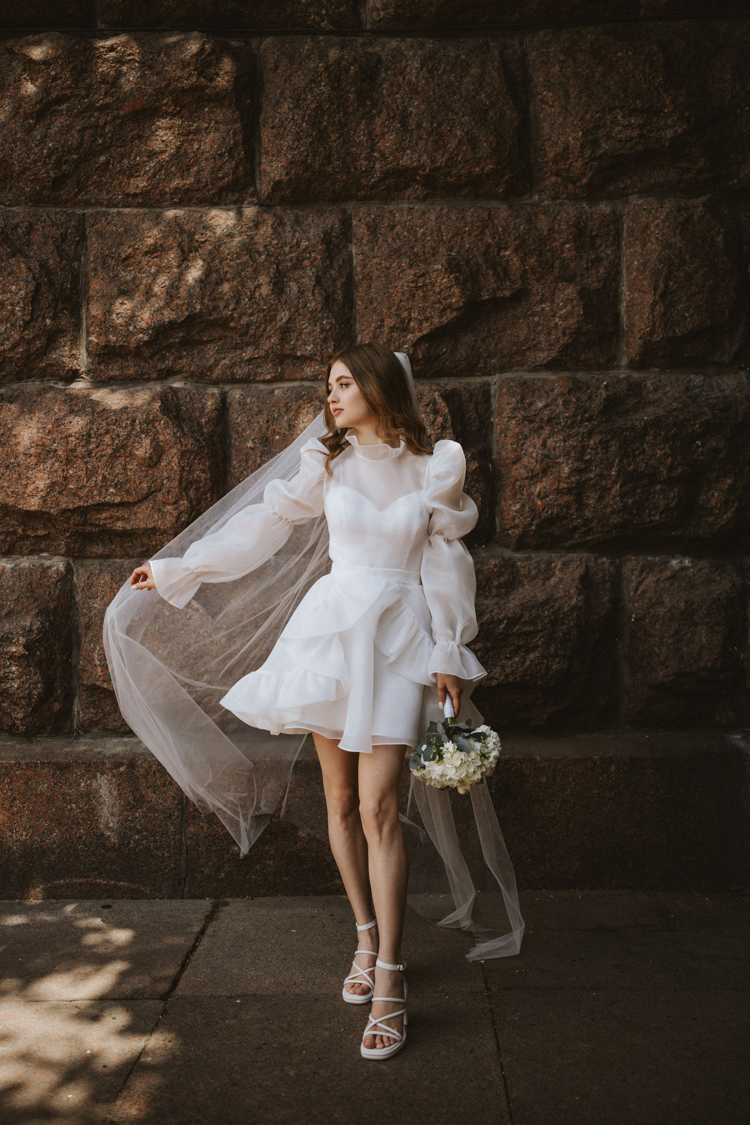 “Una sposa in un moderno mini abito bianco con maniche a sbuffo e un velo fluente si erge fiduciosa contro un muro di pietra rustico. Tiene in mano un piccolo bouquet di fiori bianchi e la brezza leggera solleva il velo, creando un effetto elegante e giocoso. La giornata luminosa e soleggiata aggiunge una calda luce alla scena”.
