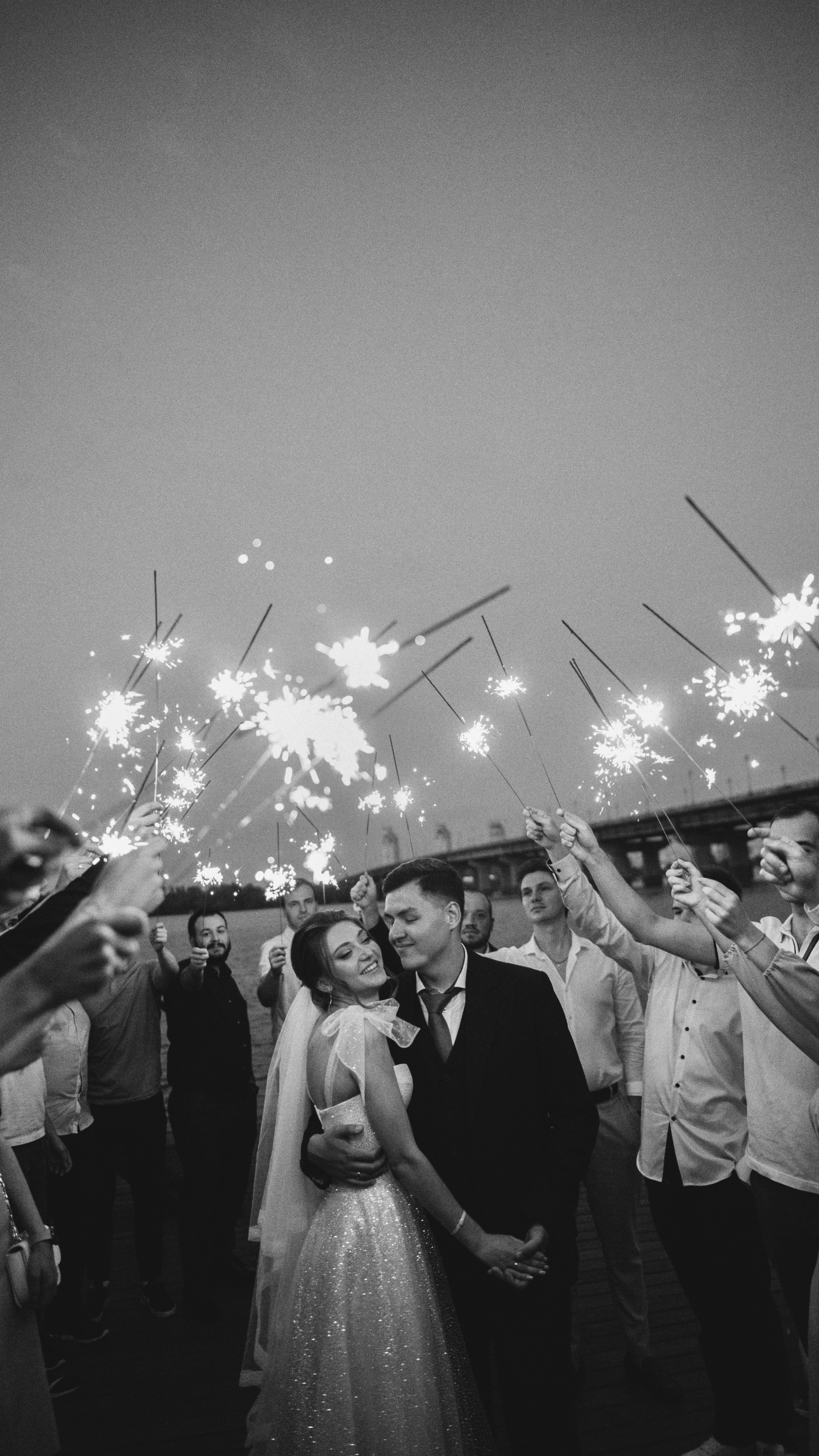 “Una foto in bianco e nero di una coppia di sposi in piedi, circondata dai loro ospiti con le stelle filanti durante la celebrazione del loro matrimonio. La coppia condivide un momento romantico, sorridendo mentre si abbraccia, con le scintille che illuminano l'atmosfera gioiosa”.