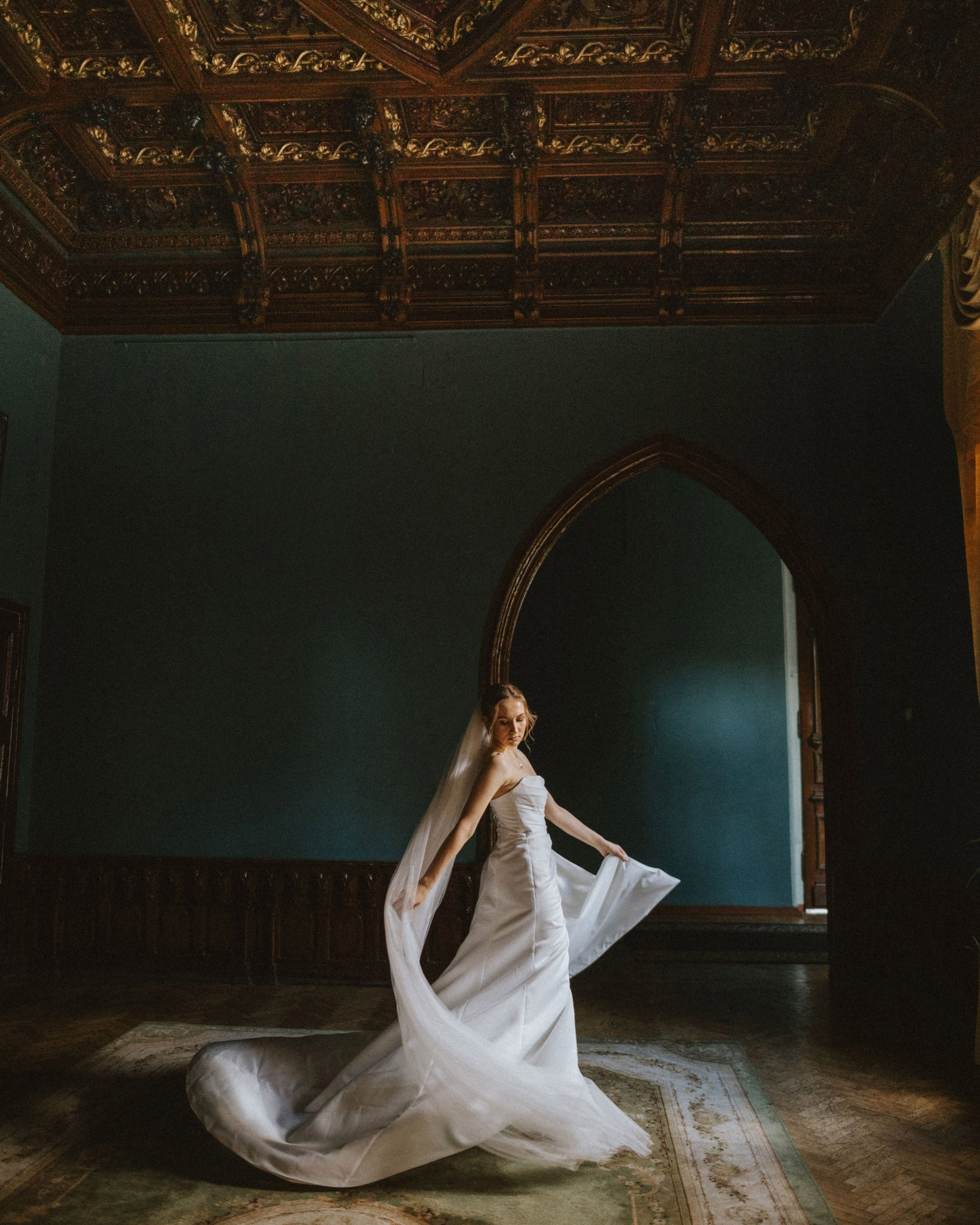 “The bride in an elegant wedding dress in an antique interior in Tuscany, her veil flowing, with a beautiful ceiling and arch in the background.”