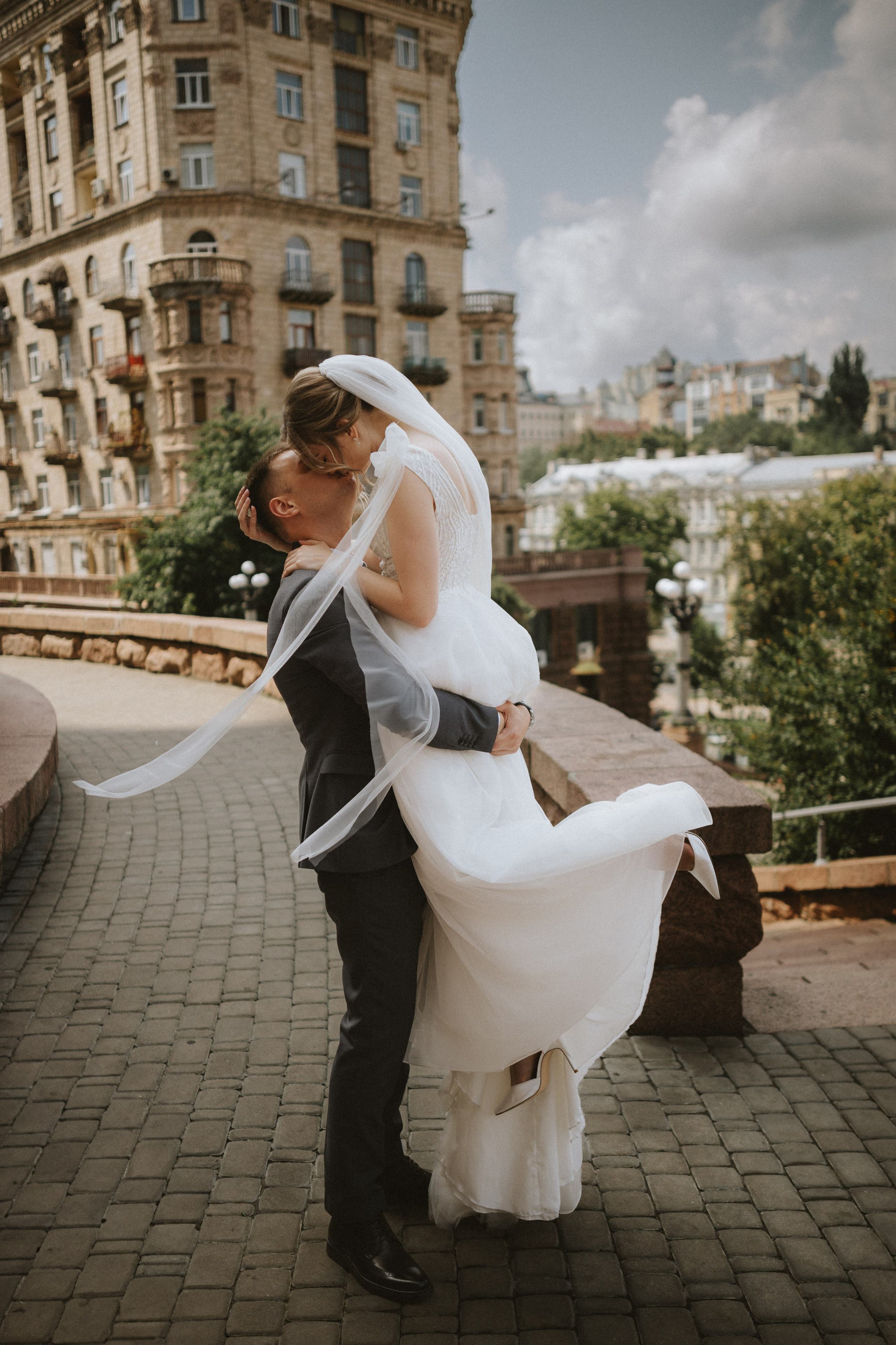 “Un momento gioioso in cui gli sposi si scambiano un bacio, con la sposa sollevata tra le braccia dello sposo. Il bellissimo sfondo della città e il velo fluente aggiungono un'atmosfera romantica, catturando l'essenza del loro amore nel loro giorno speciale.”