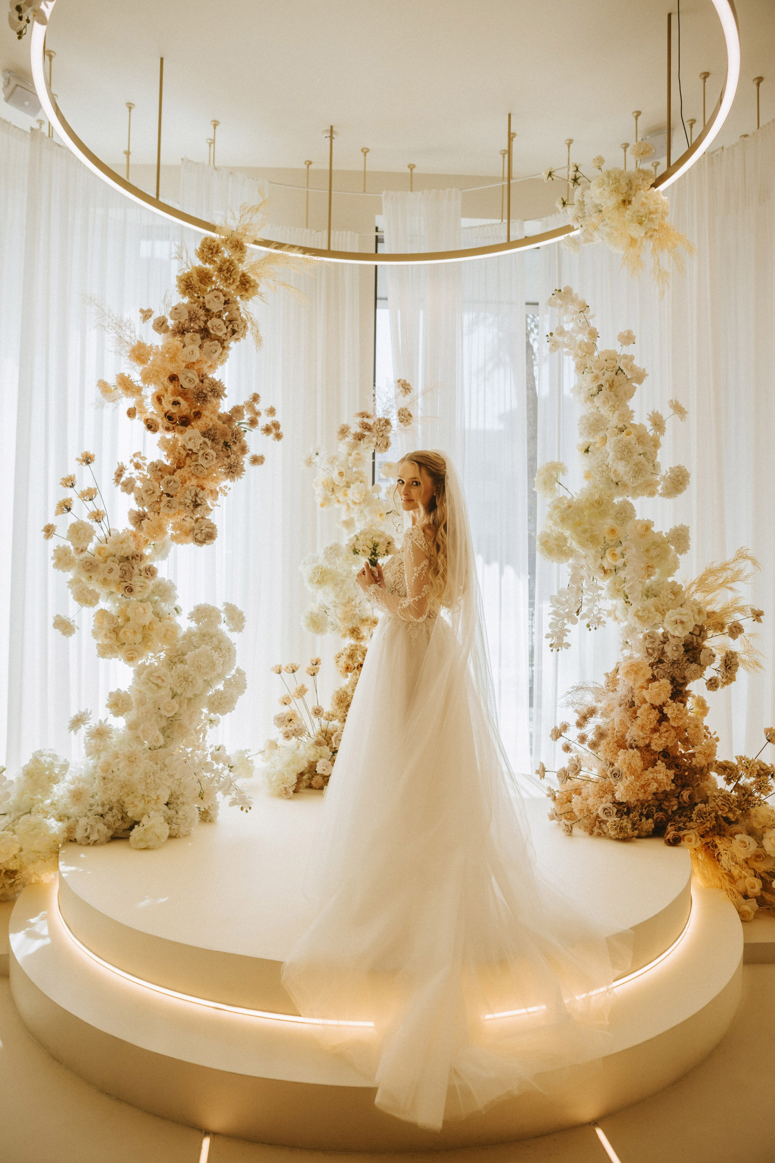 “Una sposa si erge con grazia al centro di una splendida composizione floreale, circondata da alti e delicati pilastri di fiori nelle morbide tonalità del bianco, del crema e del beige. Tiene in mano un piccolo bouquet e guarda alle sue spalle con un sorriso sereno. Lo spazio moderno e minimalista è illuminato da una lampada circolare luminosa, che accentua l'eleganza e la bellezza del momento. Il suo abito morbido e fluente aggiunge un'atmosfera eterea alla scena”.