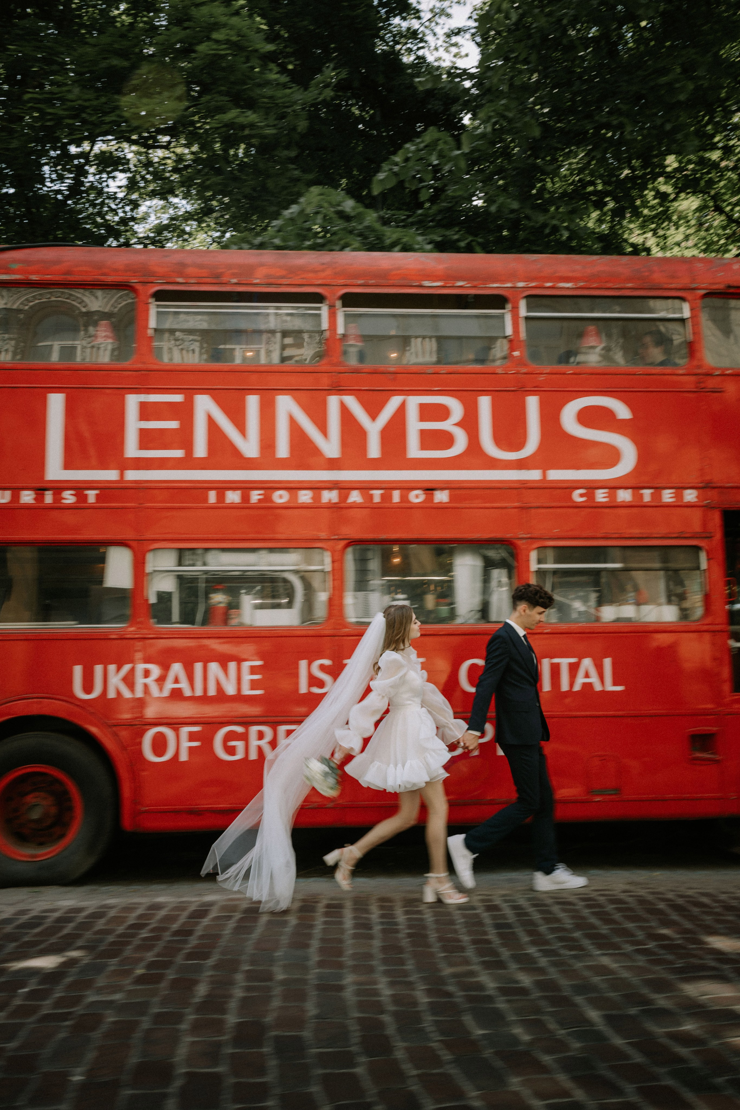 “Una coppia cammina mano nella mano, irradiando gioia, davanti a un autobus a due piani rosso brillante. La sposa, vestita con un allegro abito bianco e un velo fluente, è felice, mentre lo sposo, in abito scuro, cammina accanto a lei. L'autobus, con la scritta “LENNYBUS” e “L'UCRAINA È LA CAPITALE DEI GRANDI POPOLI”, aggiunge uno sfondo unico e vivace a questo momento speciale”.