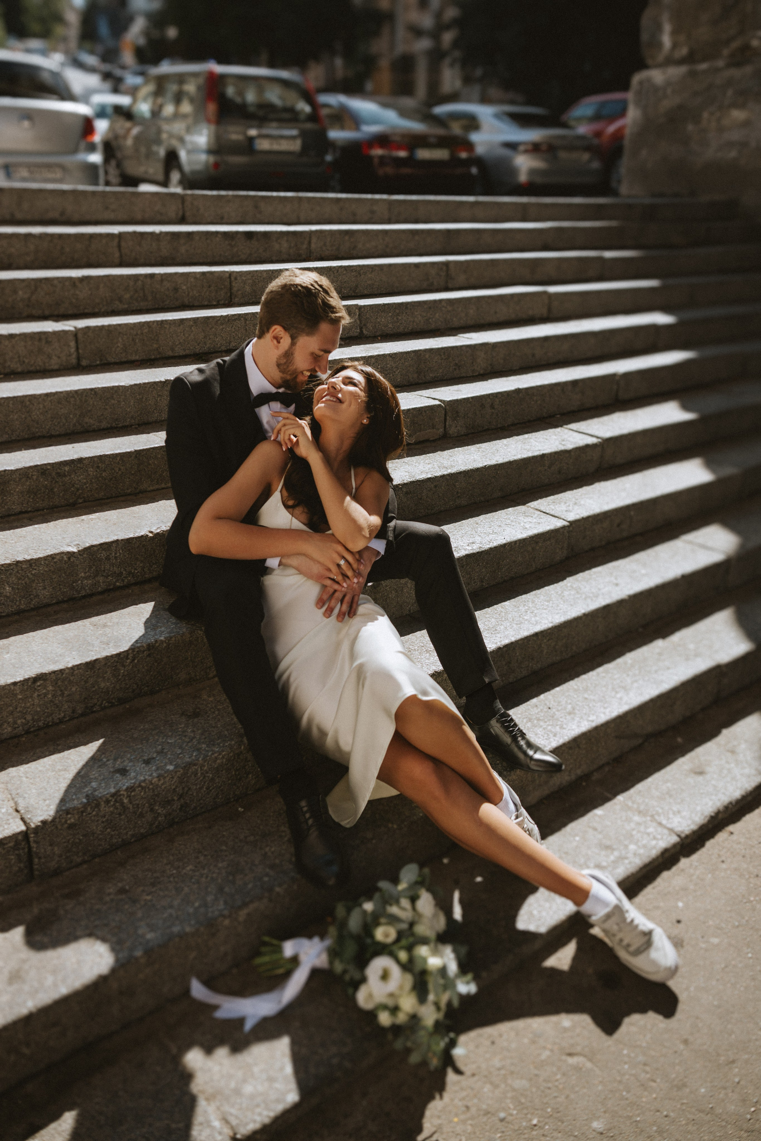 “Una sposa e uno sposo condividono un momento di gioia seduti insieme su una scala. La sposa, vestita con un elegante abito bianco abbinato a scarpe da ginnastica, ride mentre lo sposo, in smoking, le tiene amorevolmente la vita. Il bouquet di lei è appoggiato a terra accanto a loro, aggiungendo un tocco morbido alla scena vivace e giocosa sotto la luce del sole”.