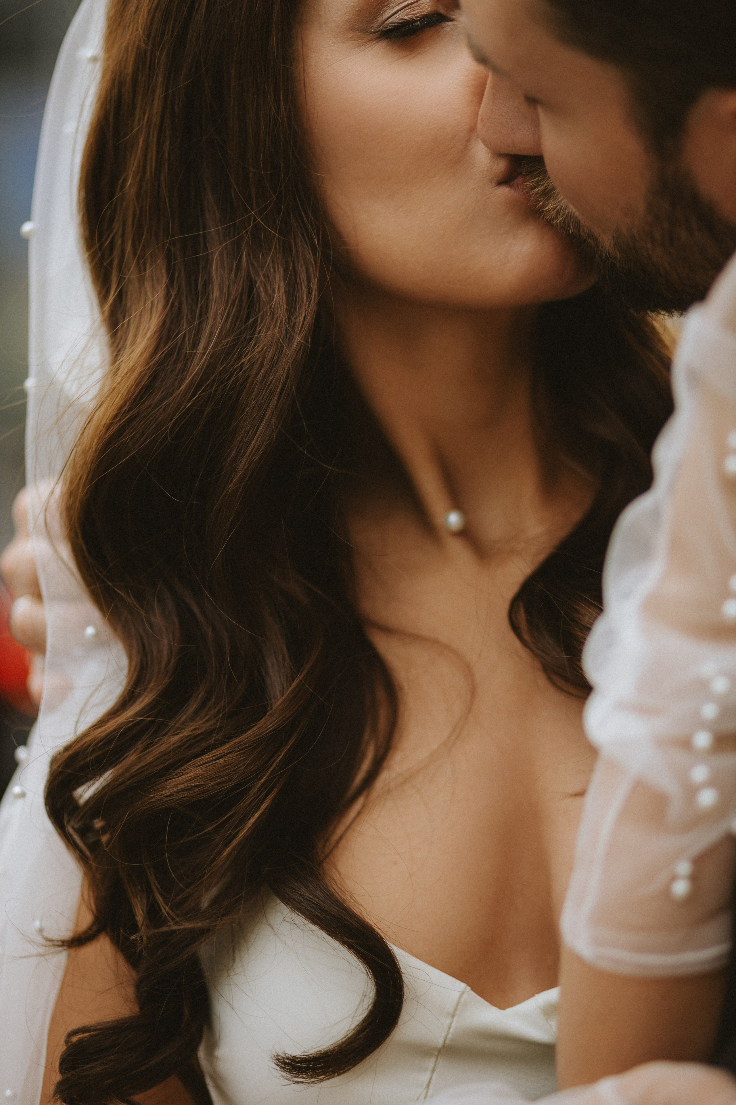 “Primo piano di una coppia che si bacia, con la sposa che indossa un velo ornato di perle e una delicata collana di perle, mentre lo sposo la abbraccia dolcemente”.