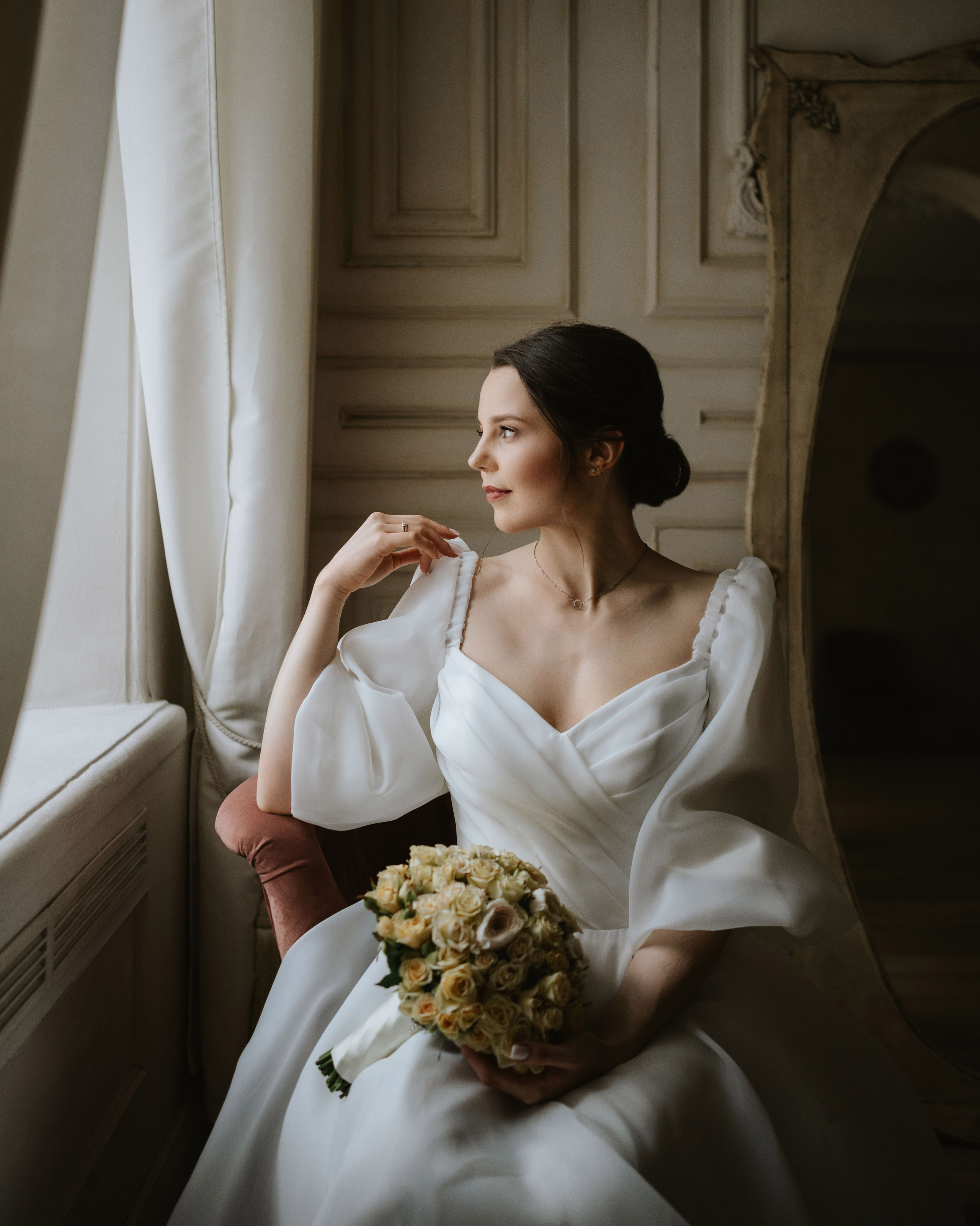 “The bride with a bouquet of roses, sitting in a historic interior with windows, a wedding in one of Italy’s romantic cities, such as Verona or Florence.”