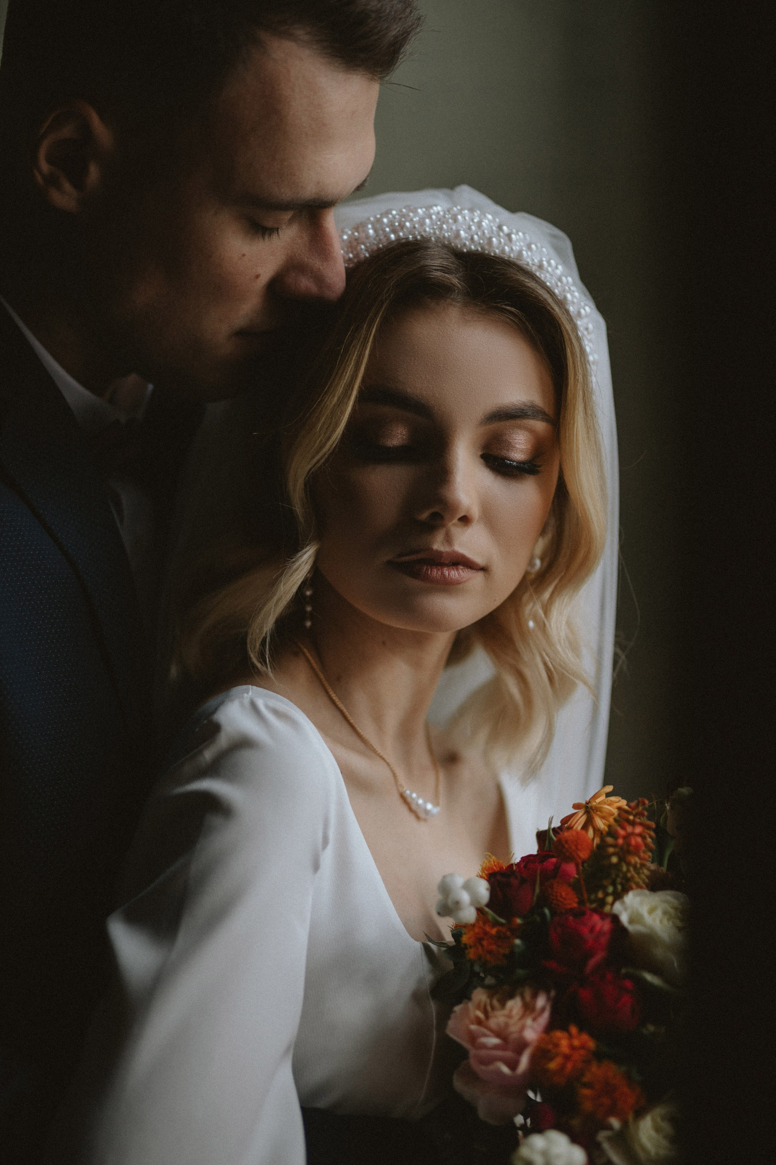 “Fotografo di matrimonio che cattura un momento intimo tra gli sposi, con la sposa che tiene in mano un bouquet di fiori vivaci. La luce soffusa mette in risalto la sua collana di perle e il velo, mentre lo sposo la abbraccia dolcemente, creando un'atmosfera romantica, ideale per un matrimonio in Toscana.”