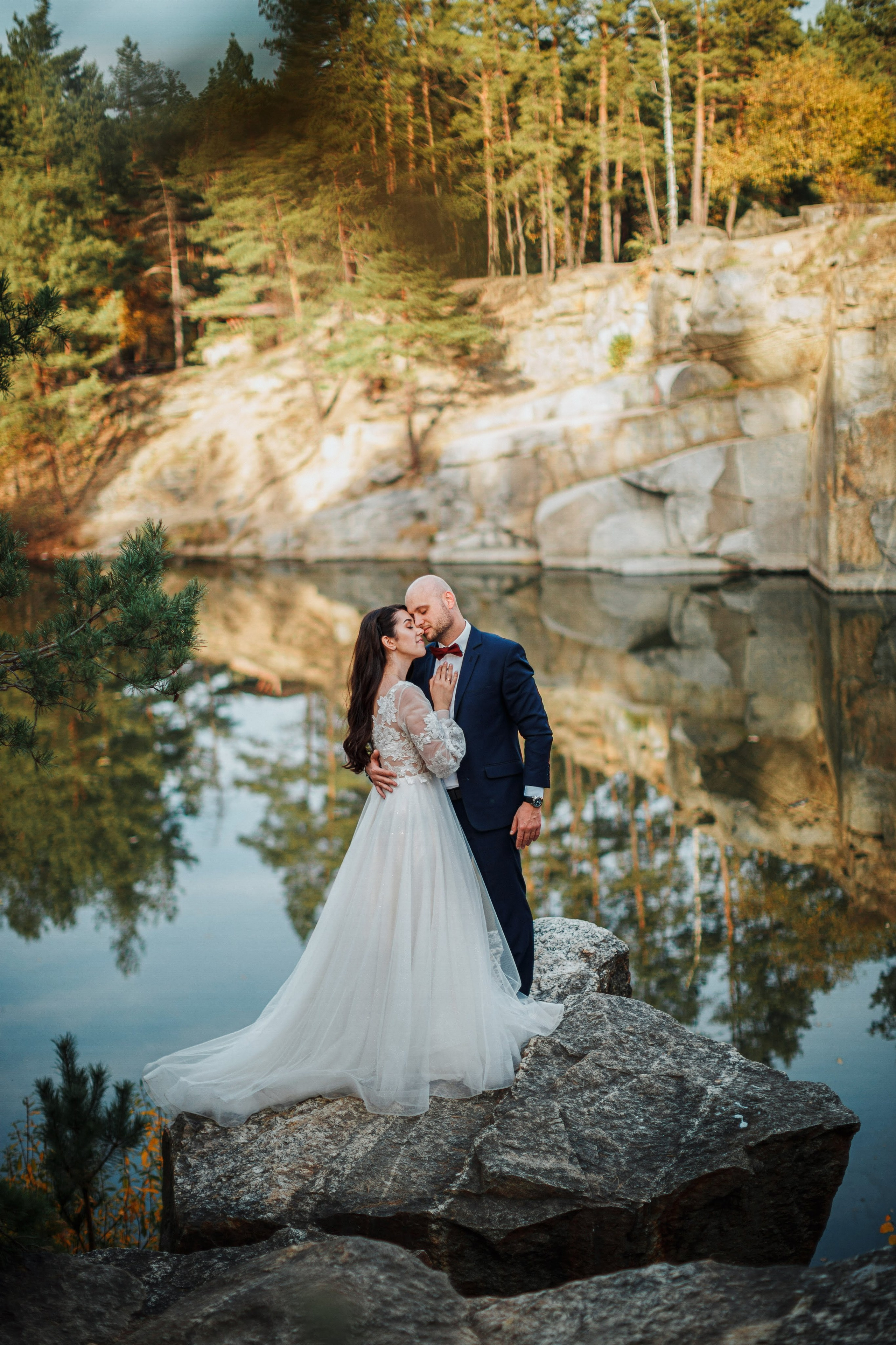 “Una sposa e uno sposo si scambiano un bacio intimo in piedi su una grande roccia in riva a un lago sereno, circondati da alberi e natura. La sposa indossa un fluente abito bianco con intricati dettagli in pizzo, mentre lo sposo indossa un completo blu con papillon rosso. La scena di pace si riflette nell'acqua calma”.