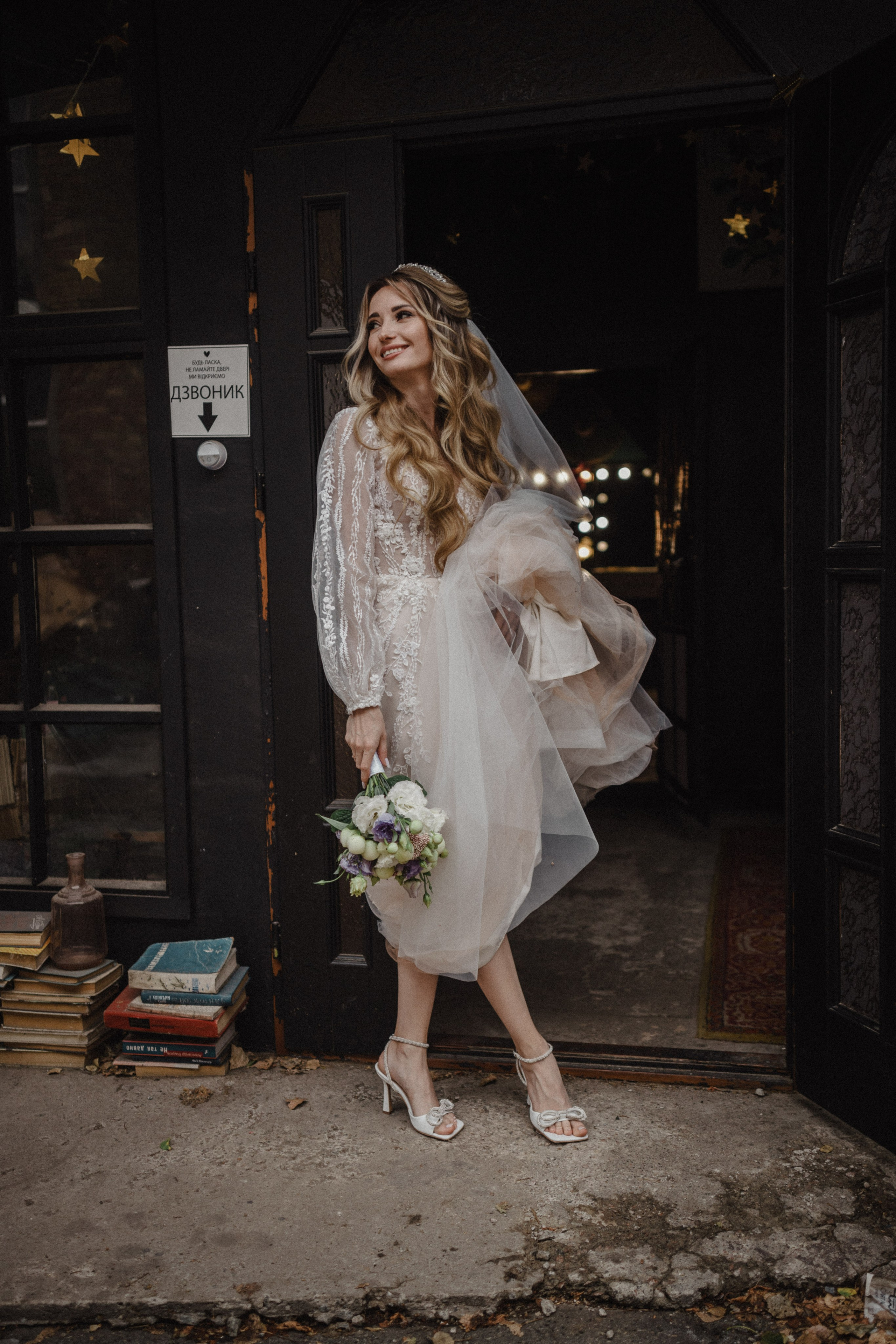 “Una sposa che indossa un delicato abito trasparente con intricati dettagli in pizzo si trova all'ingresso di un affascinante edificio. Tiene in mano un bouquet di fiori bianchi e viola, sorride dolcemente mentre si aggiusta delicatamente il velo, emanando un'aura gioiosa ed elegante. La scena è incorniciata dal caldo bagliore delle luci sullo sfondo e dai libri d'epoca impilati ai suoi piedi”.