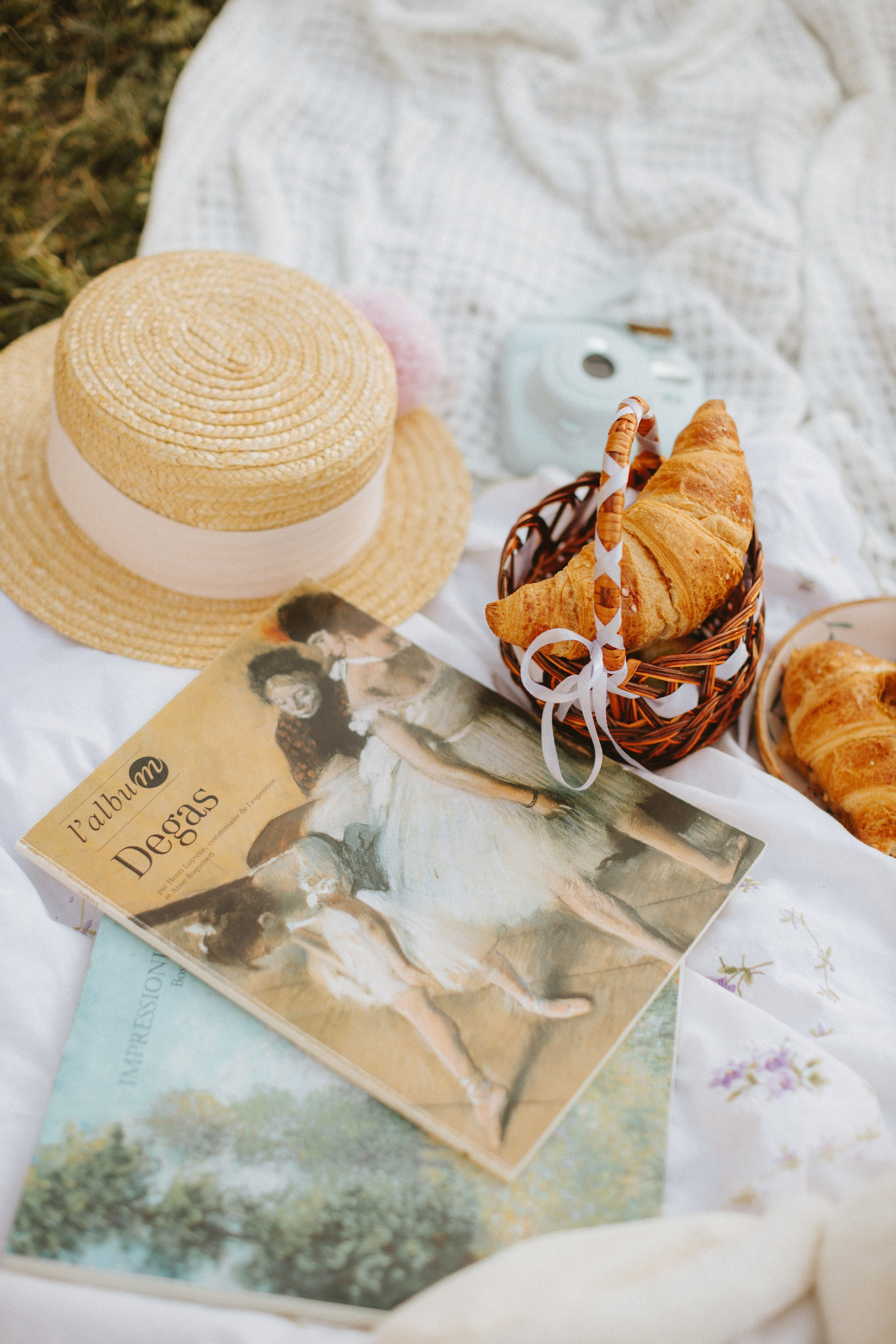 “Una serena scena di picnic con un cappello di paglia, un cestino pieno di croissant e due libri d'arte con dipinti di Degas. Una macchina fotografica è posizionata accanto agli oggetti, mentre una morbida coperta bianca e dei fiori delicati contribuiscono a creare un'atmosfera tranquilla”.
